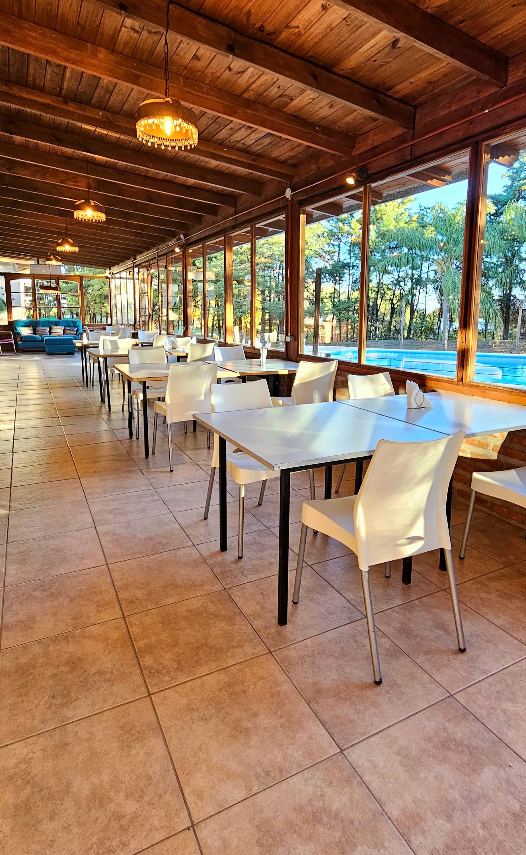 Outdoor dining area with white tables, chairs, and wood ceiling, overlooking a pool and trees.