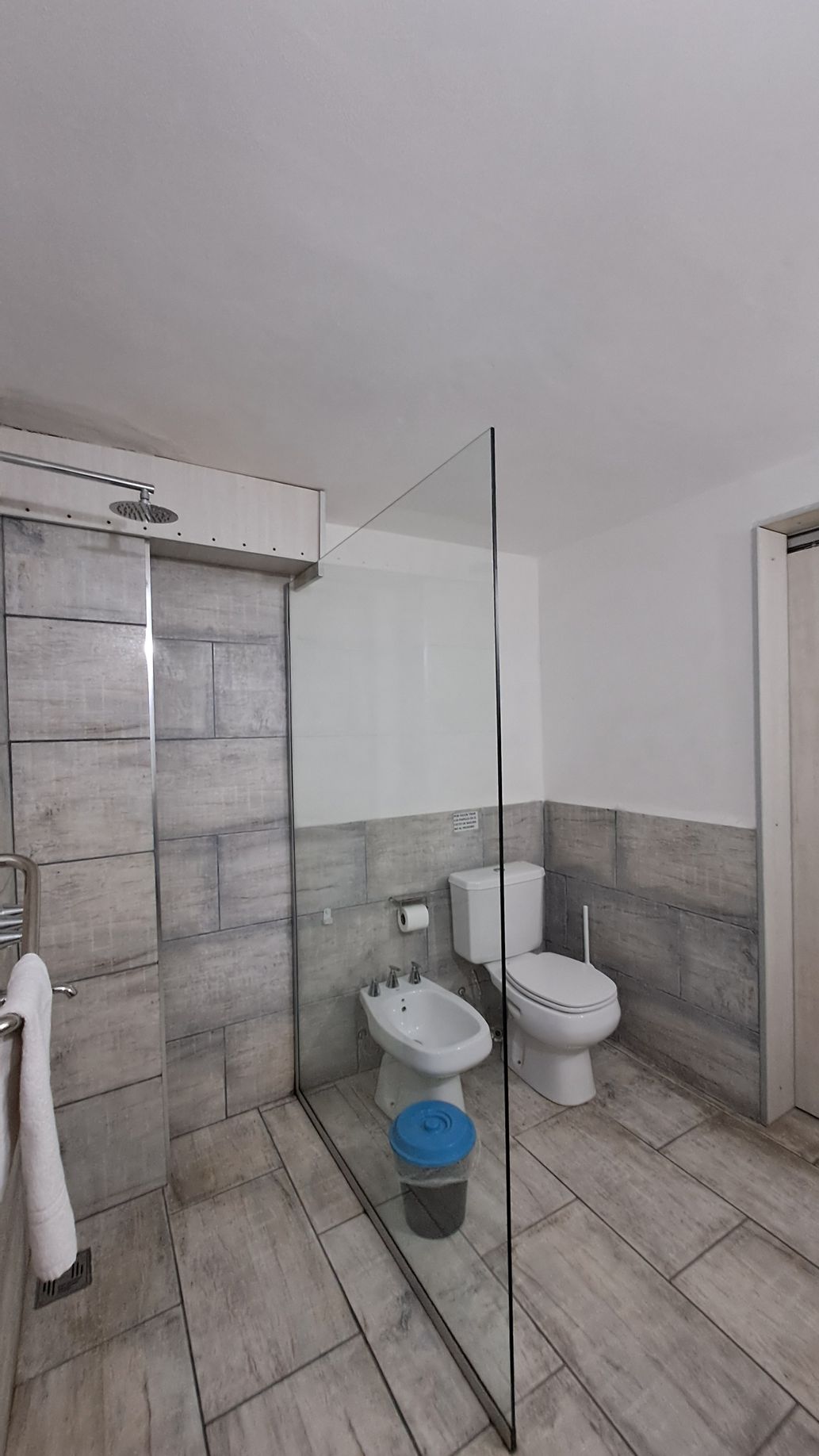 Bathroom with a walk-in shower, toilet, and bidet. Gray tiled walls and floor, white walls and a glass shower door.
