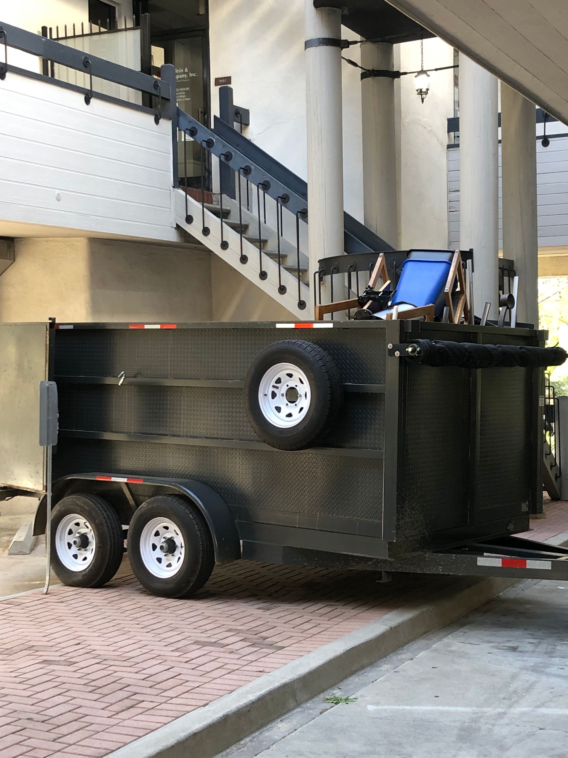 Black trailer parked on a brick-paved surface beneath a building with stairs.