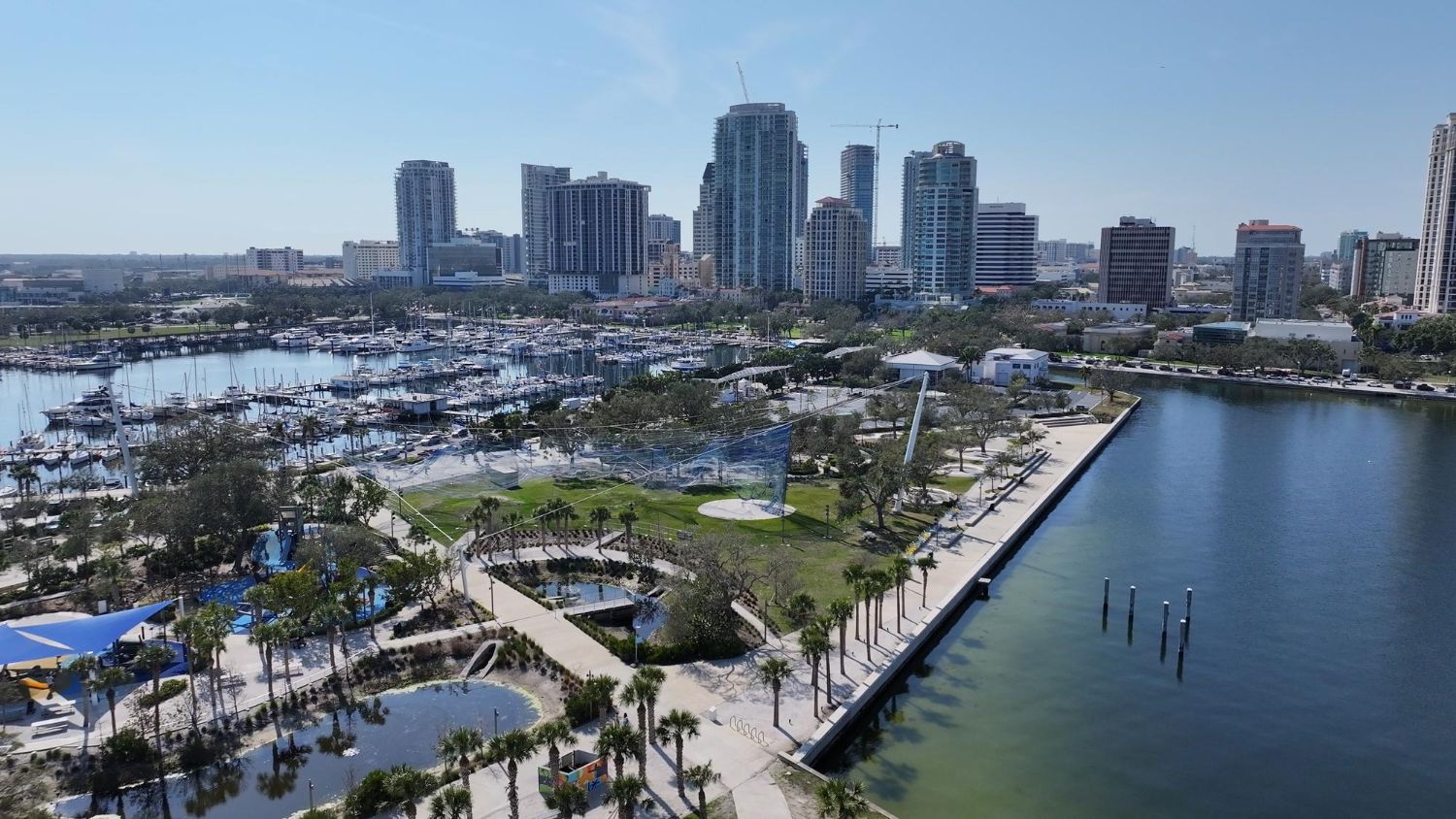 Marina waterfront with boats, palm-lined promenade, and modern high-rise skyline under a clear blue sky