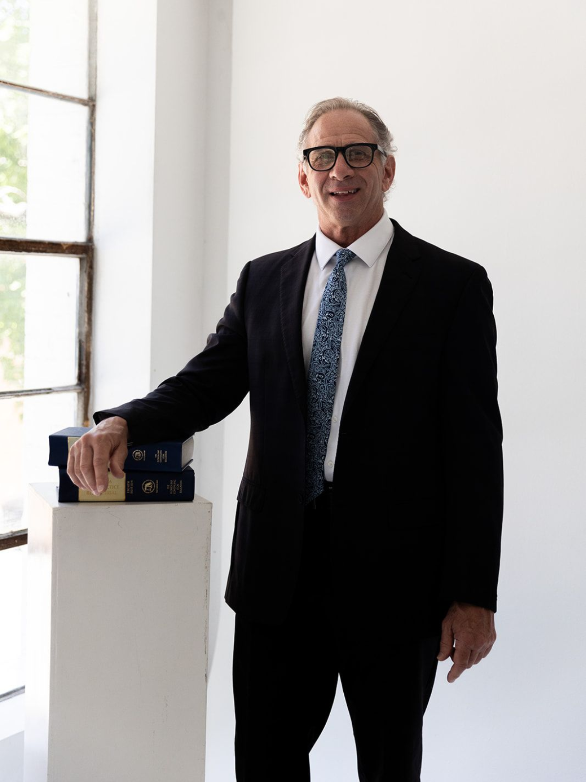 Man in a dark suit standing beside a pedestal with a book in a bright, minimalist room