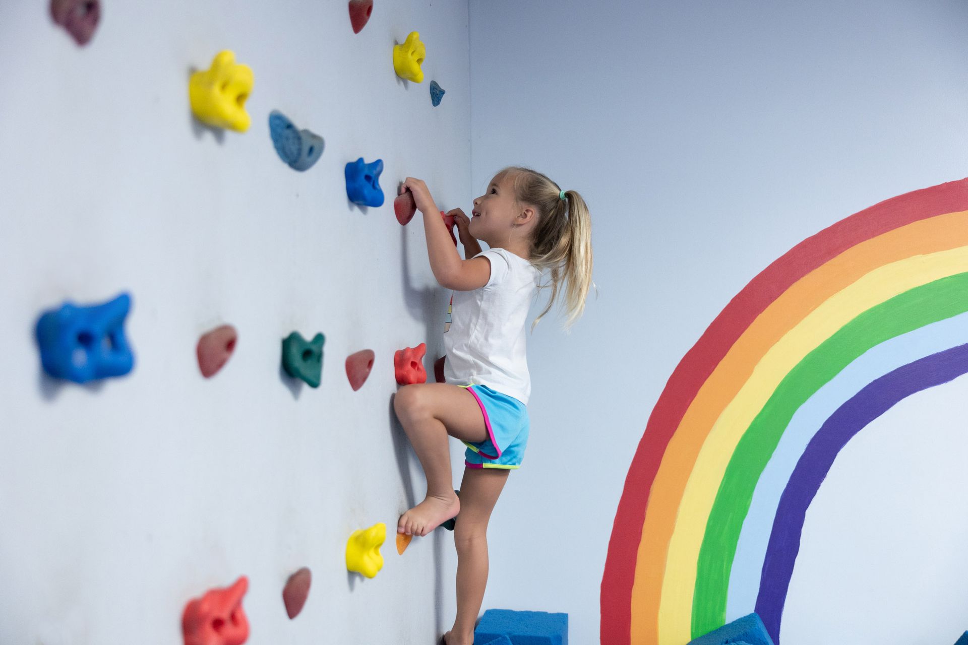 Child climbing colorful indoor rock wall beside painted rainbow mural
