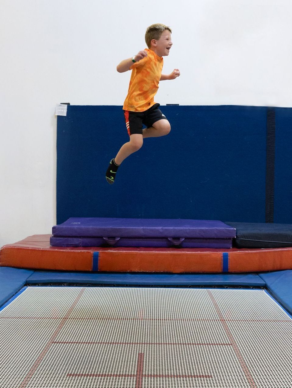 Boy in orange shirt jumping on a trampoline in an indoor gym with blue safety mats.