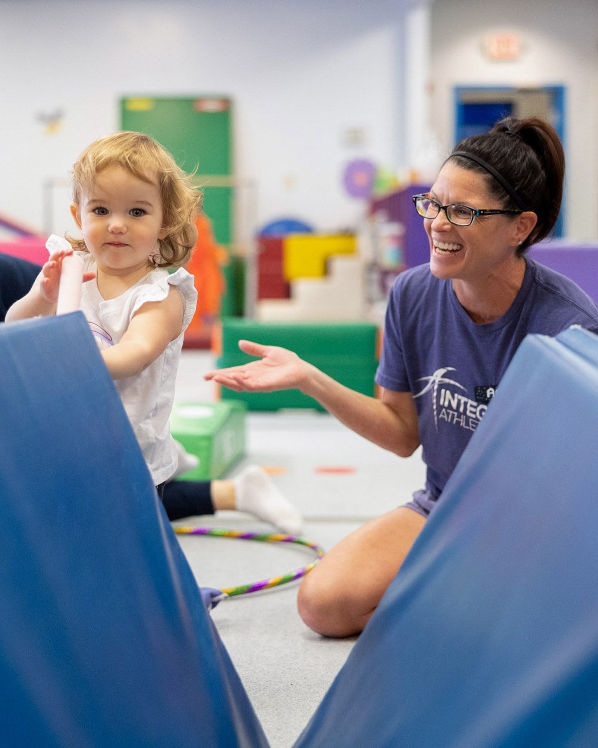 Adult and child playing in a colorful indoor gym, smiling beside blue mats.