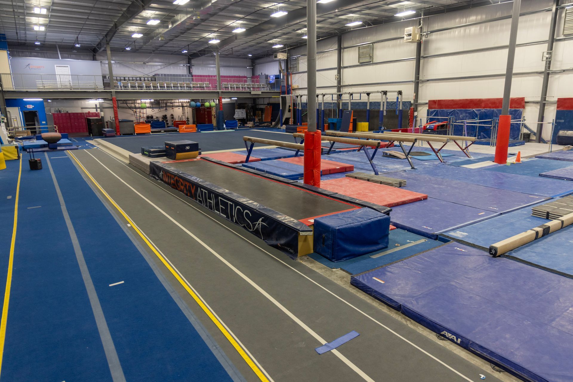 Indoor gymnastics training gym with blue mats, balance beams, and vault equipment under bright lights