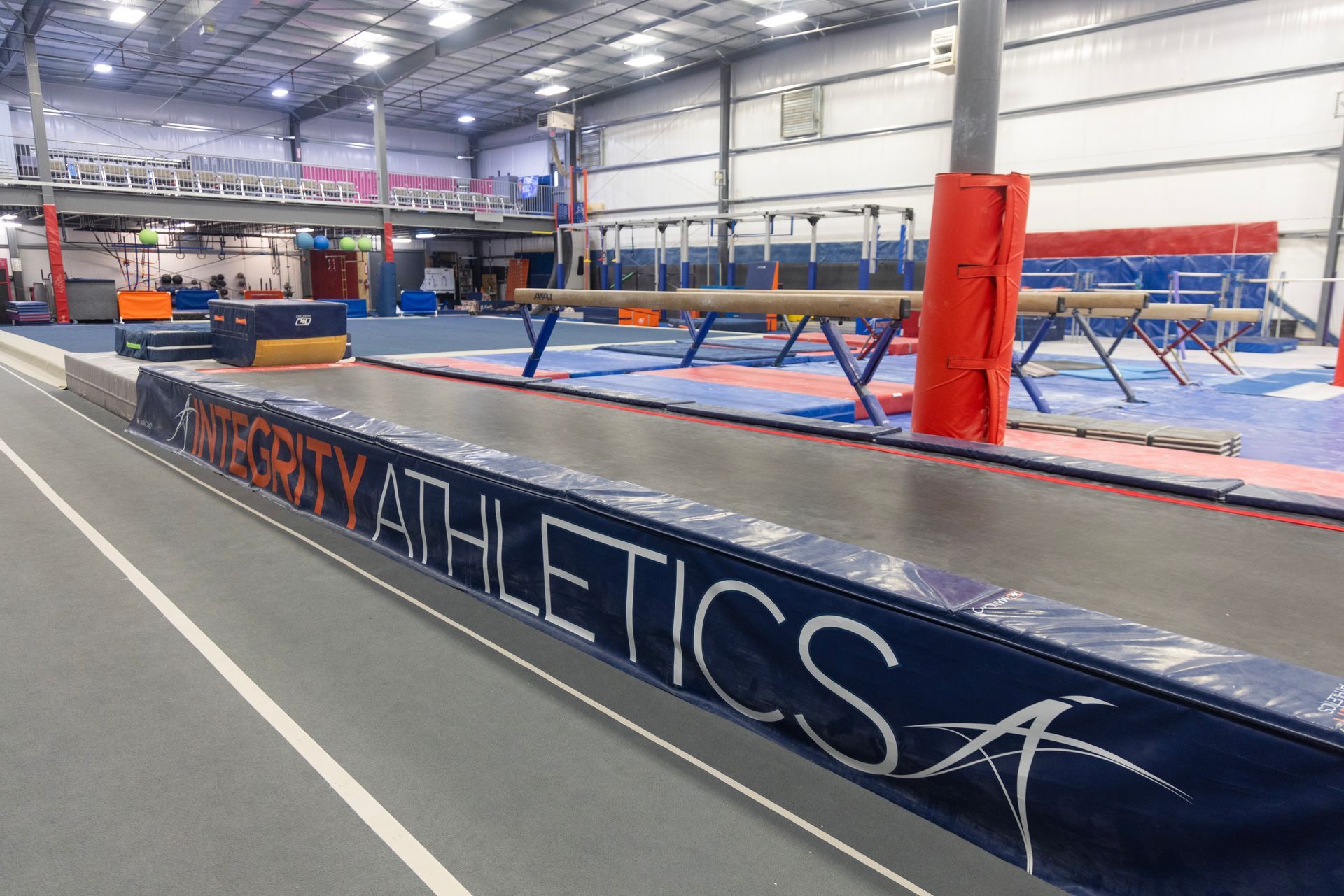 Indoor gymnastics training facility with blue mats, balance beams, and a large trampoline labeled “GYMNASTICS”