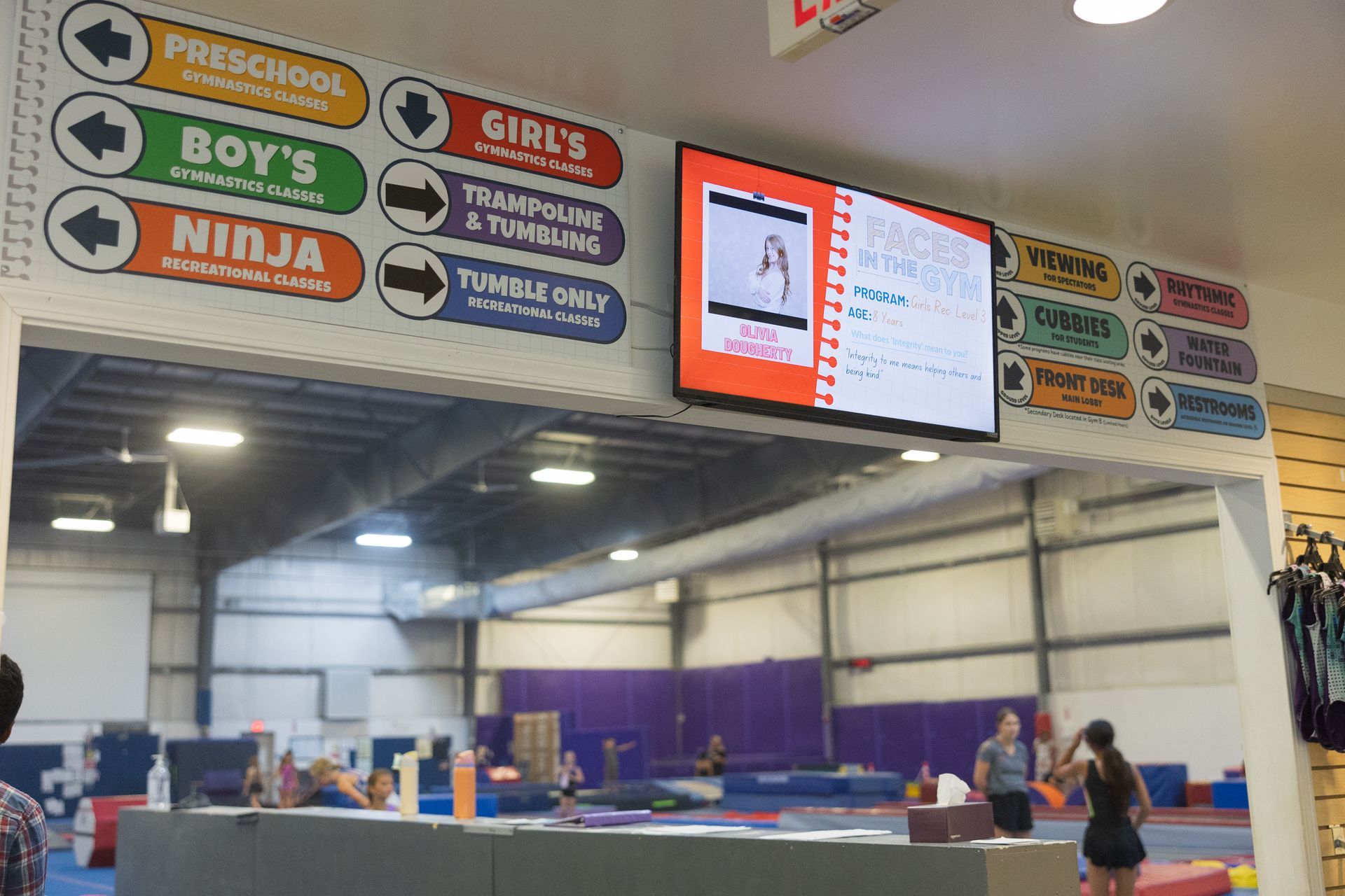 Indoor gymnastics gym with colorful directional signs and a digital scoreboard above mats