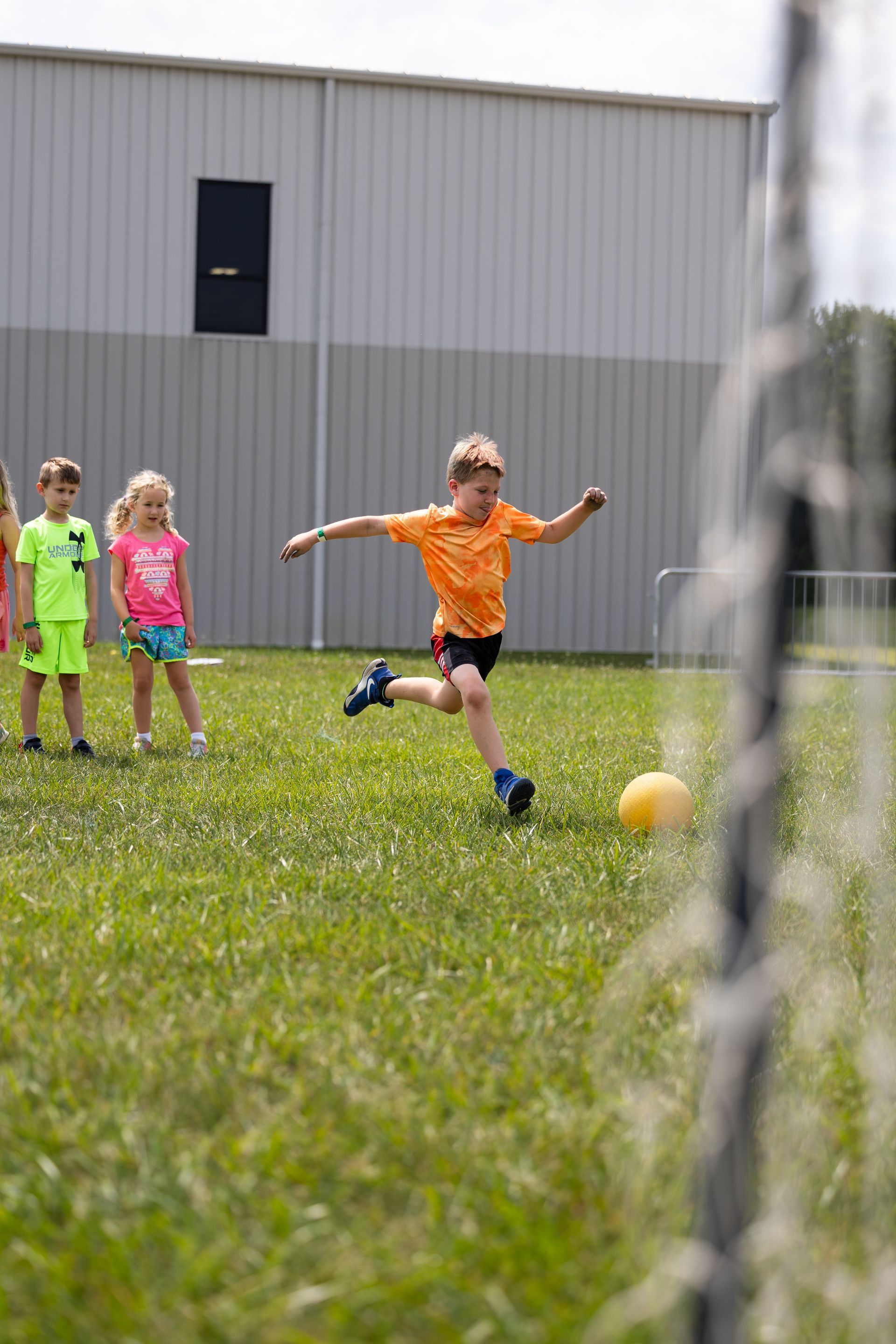 Children playing soccer on a grassy field, with one child in an orange shirt kicking the ball near a goal.