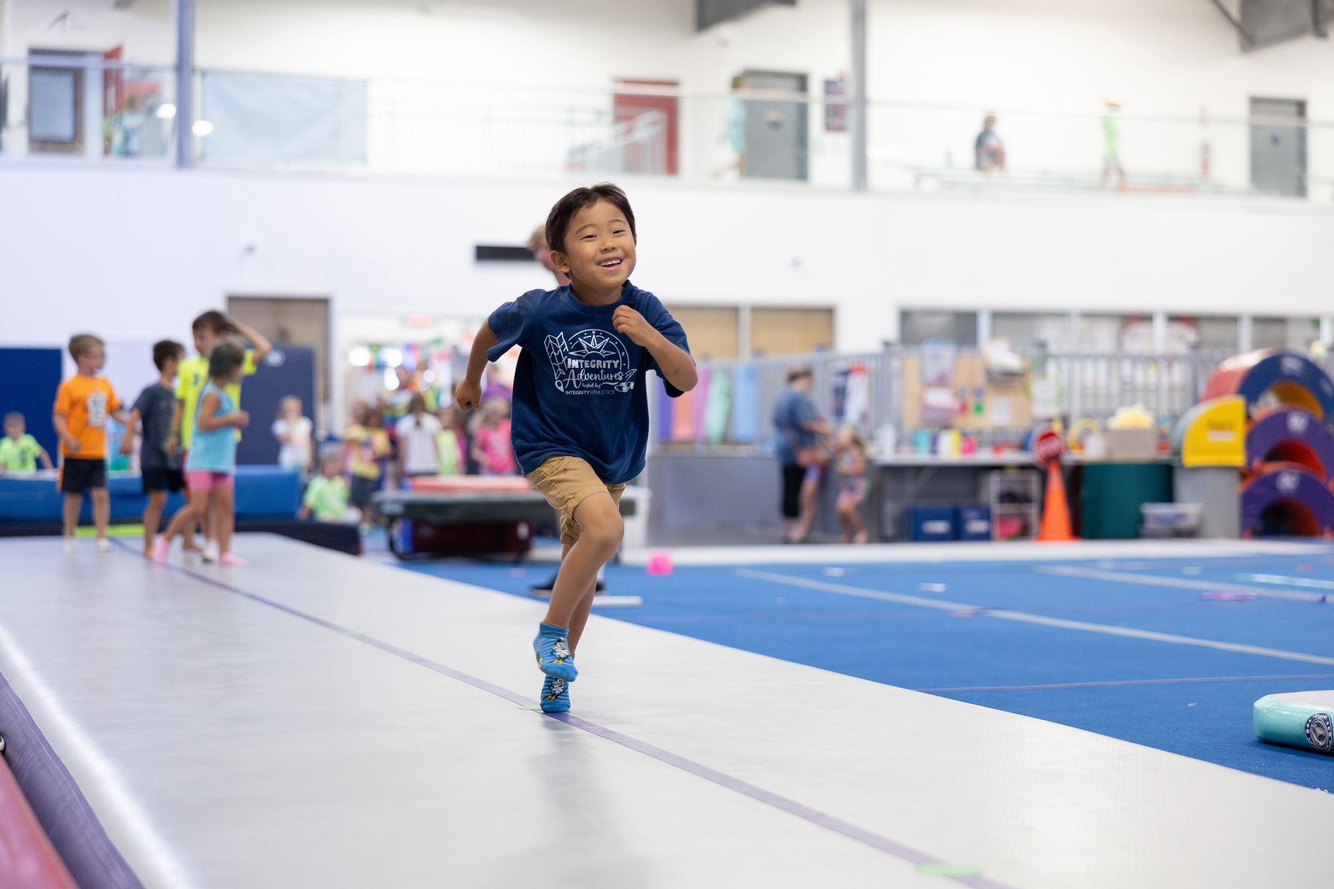 Child running on an indoor gym track with blue mats and other children in the background