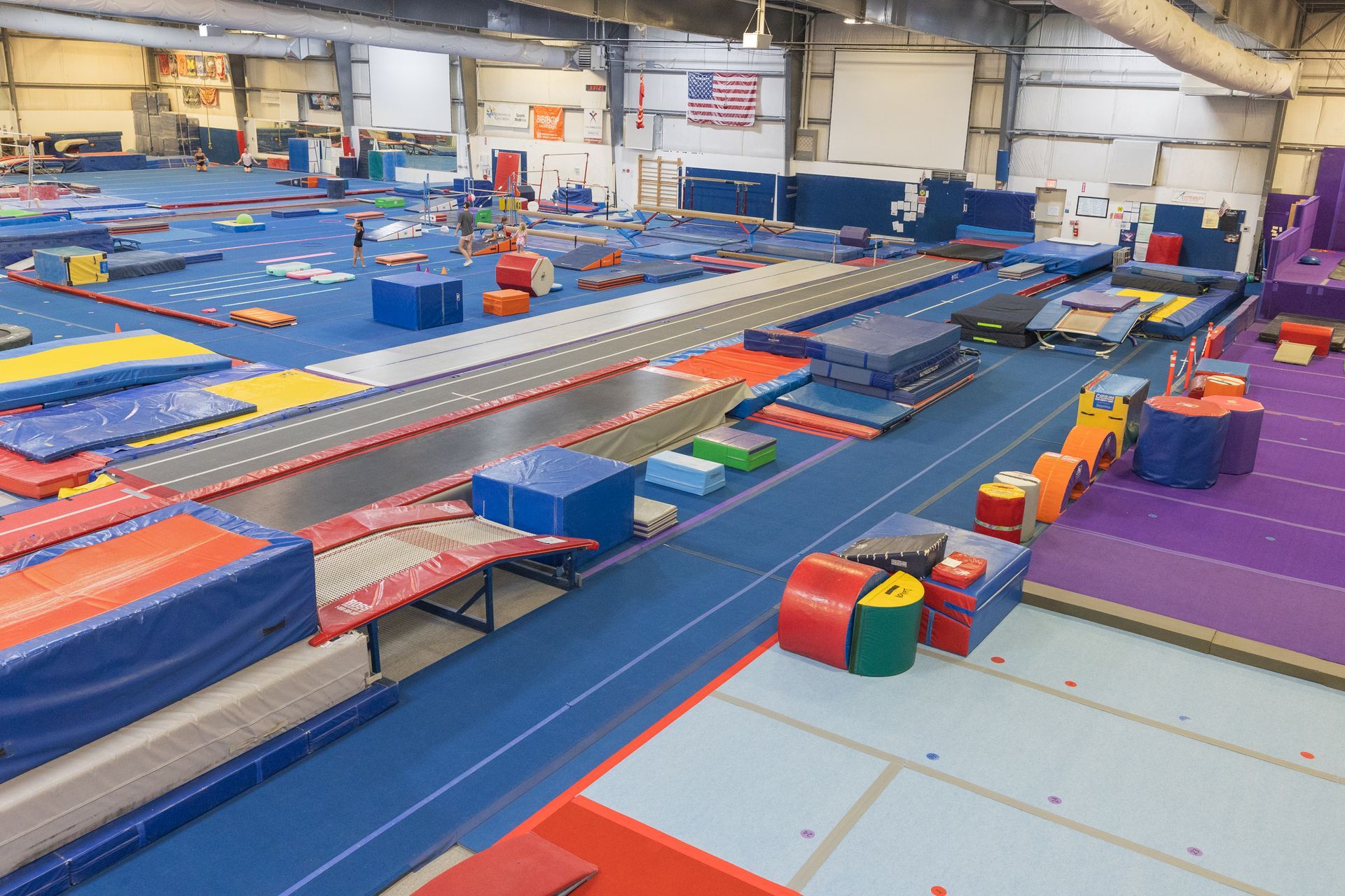 Indoor gymnastics training area with blue mats, balance beams, trampolines, and colorful equipment.