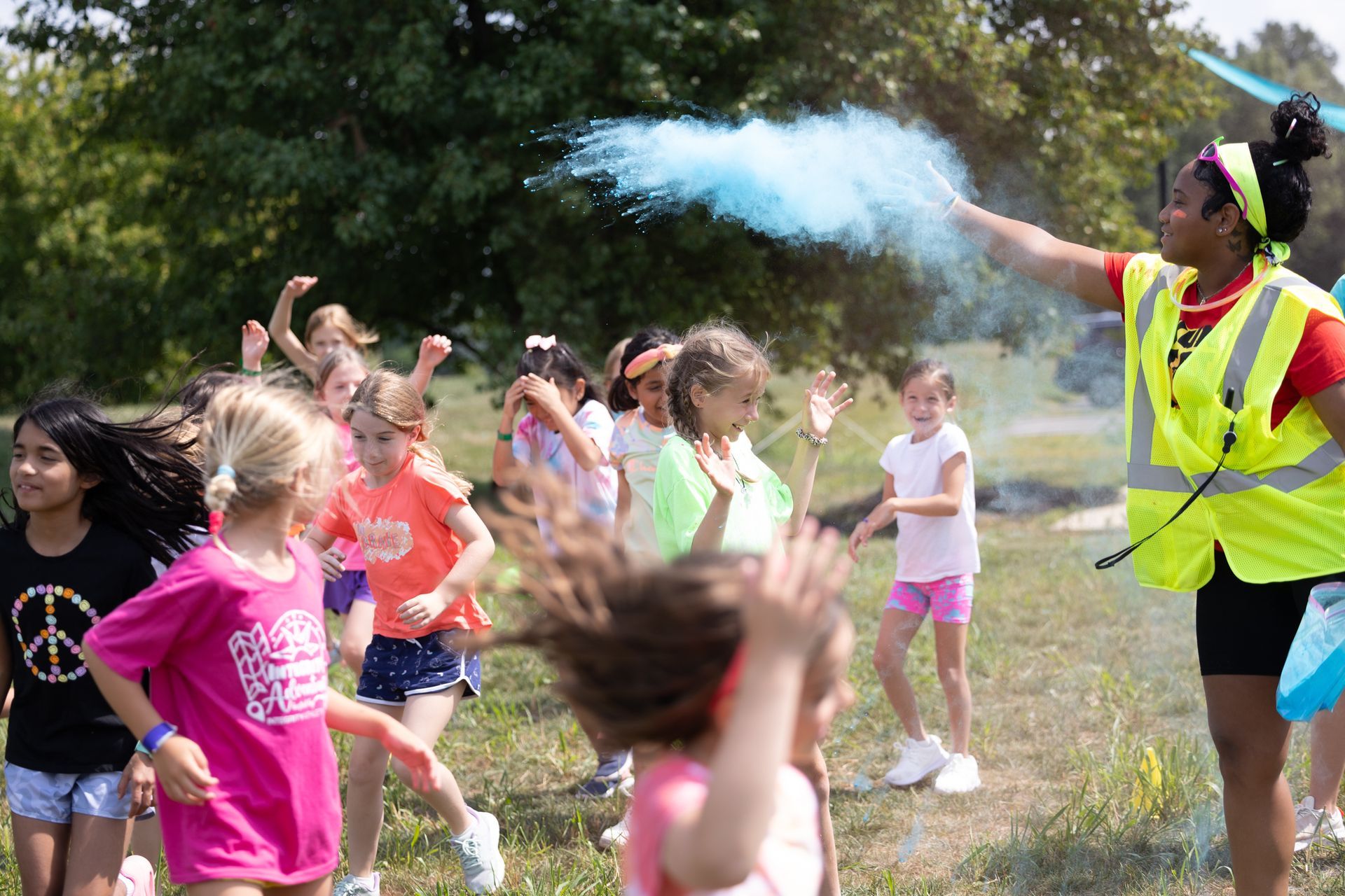 Children outdoors play in water spray from an adult in a yellow vest at a park
