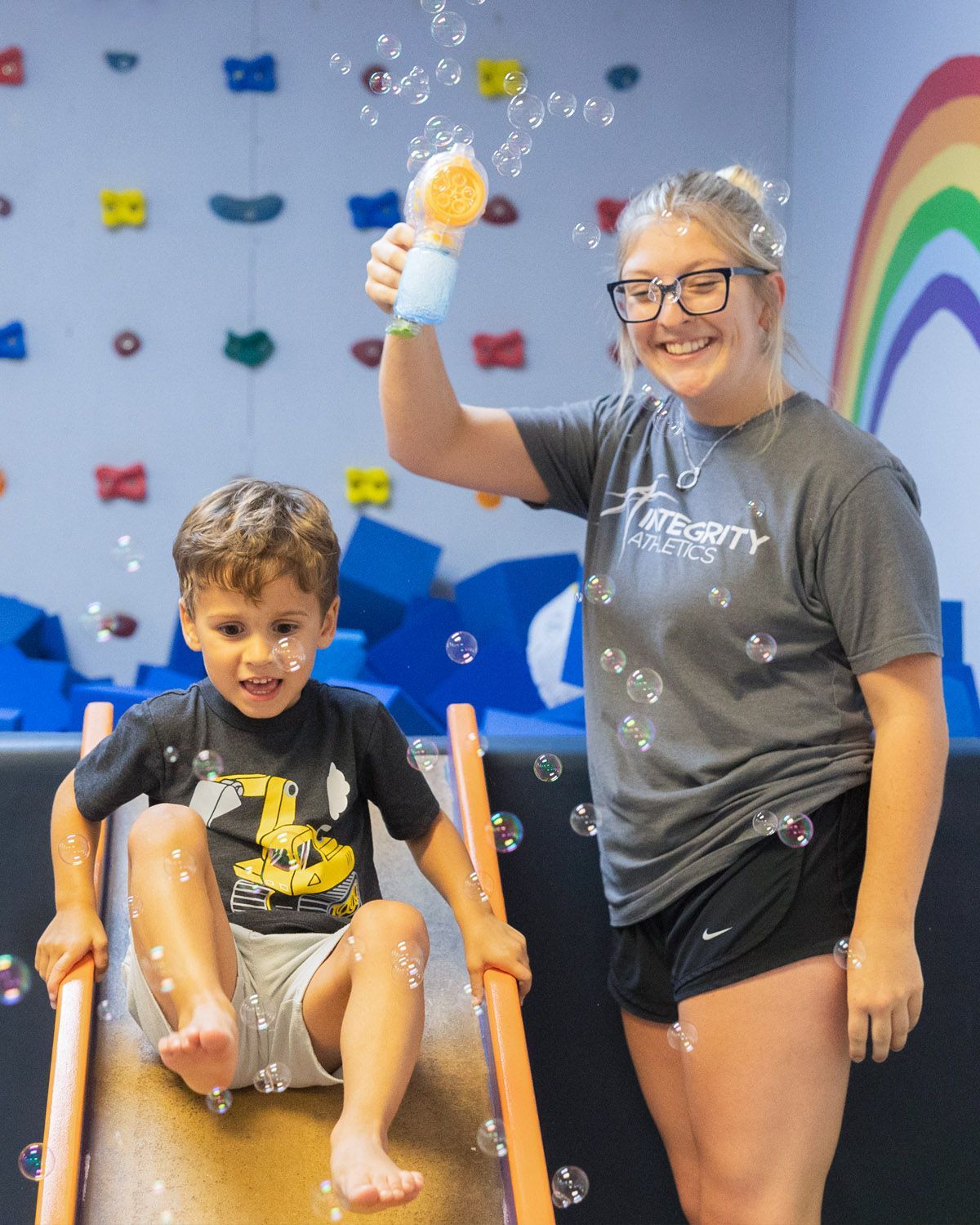 Woman and child smiling at an indoor climbing gym, holding a plush toy beside a colorful wall.