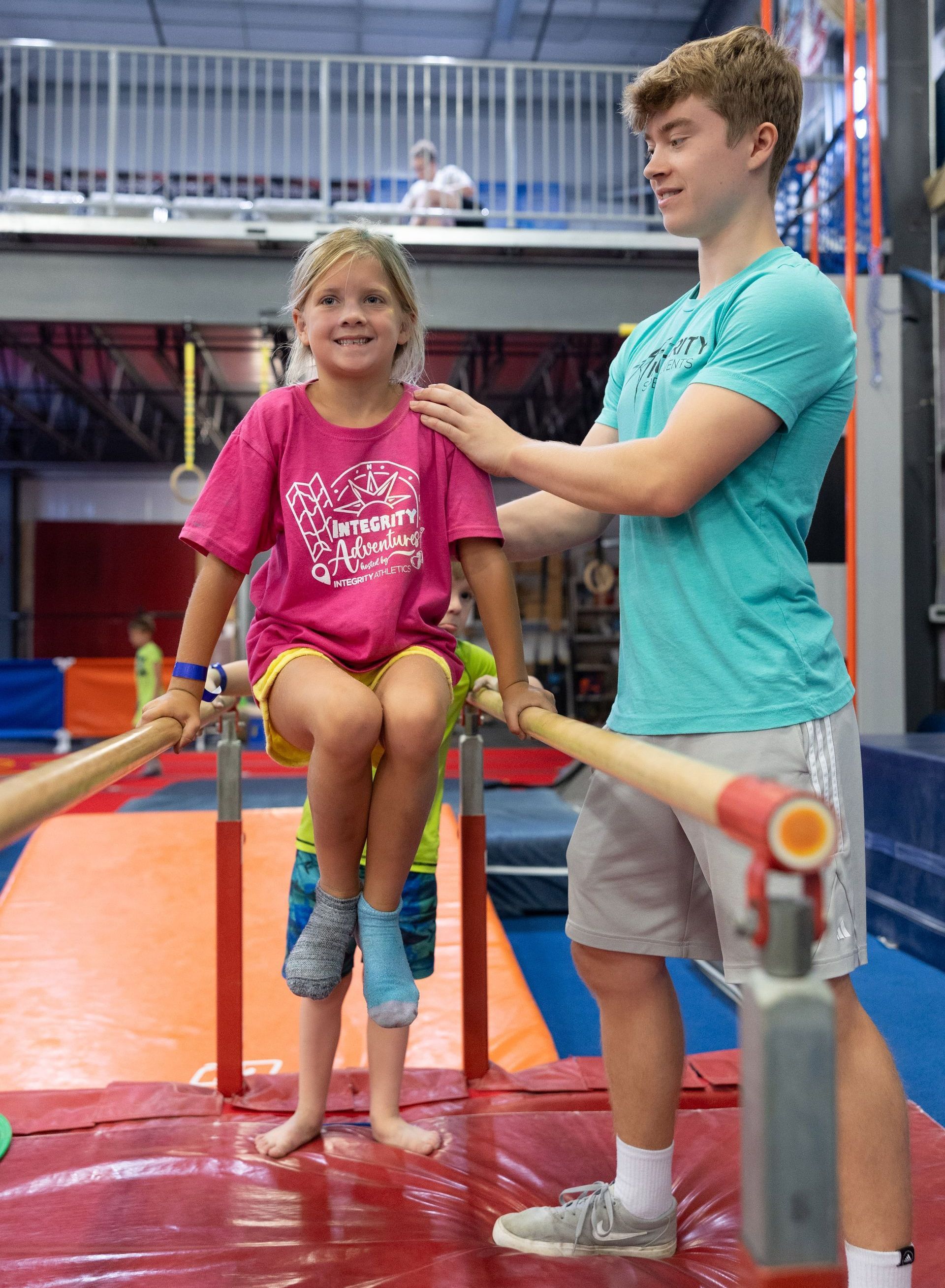 Girl on gym bars with coach spotting her indoors, wearing pink and teal in a gymnastics facility