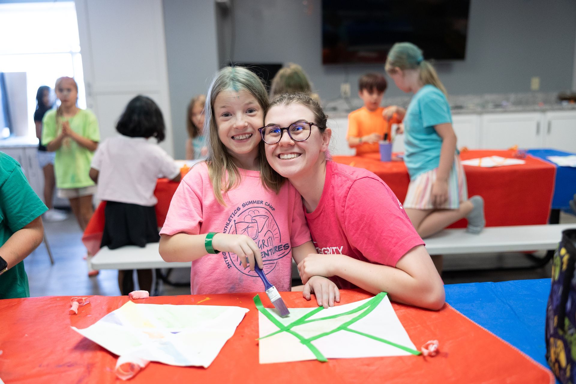 Two smiling children lean over a craft table with paper and tape at a busy indoor event.