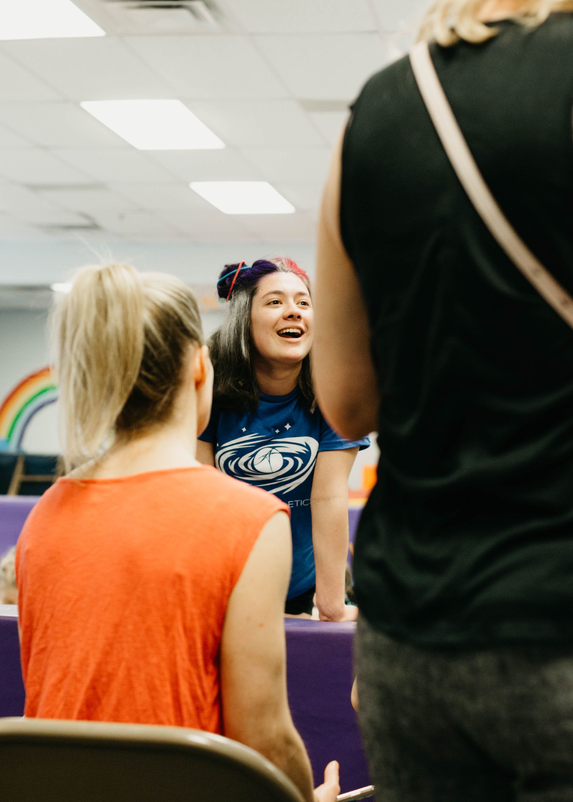 Woman speaking and smiling in a classroom, with two people seen from behind in the foreground.