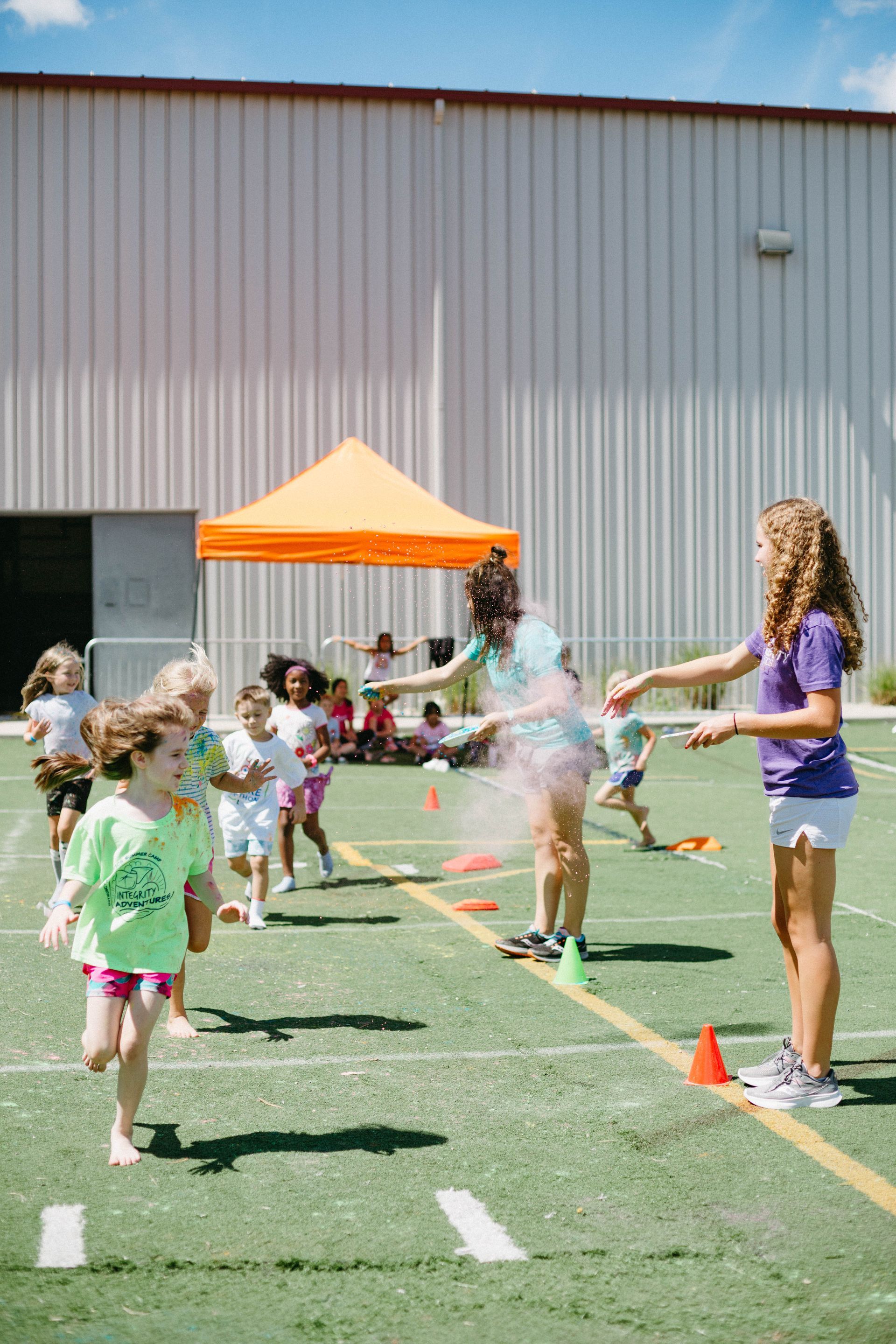 Children playing an outdoor ring toss game on a grassy field with orange cones and a building backdrop