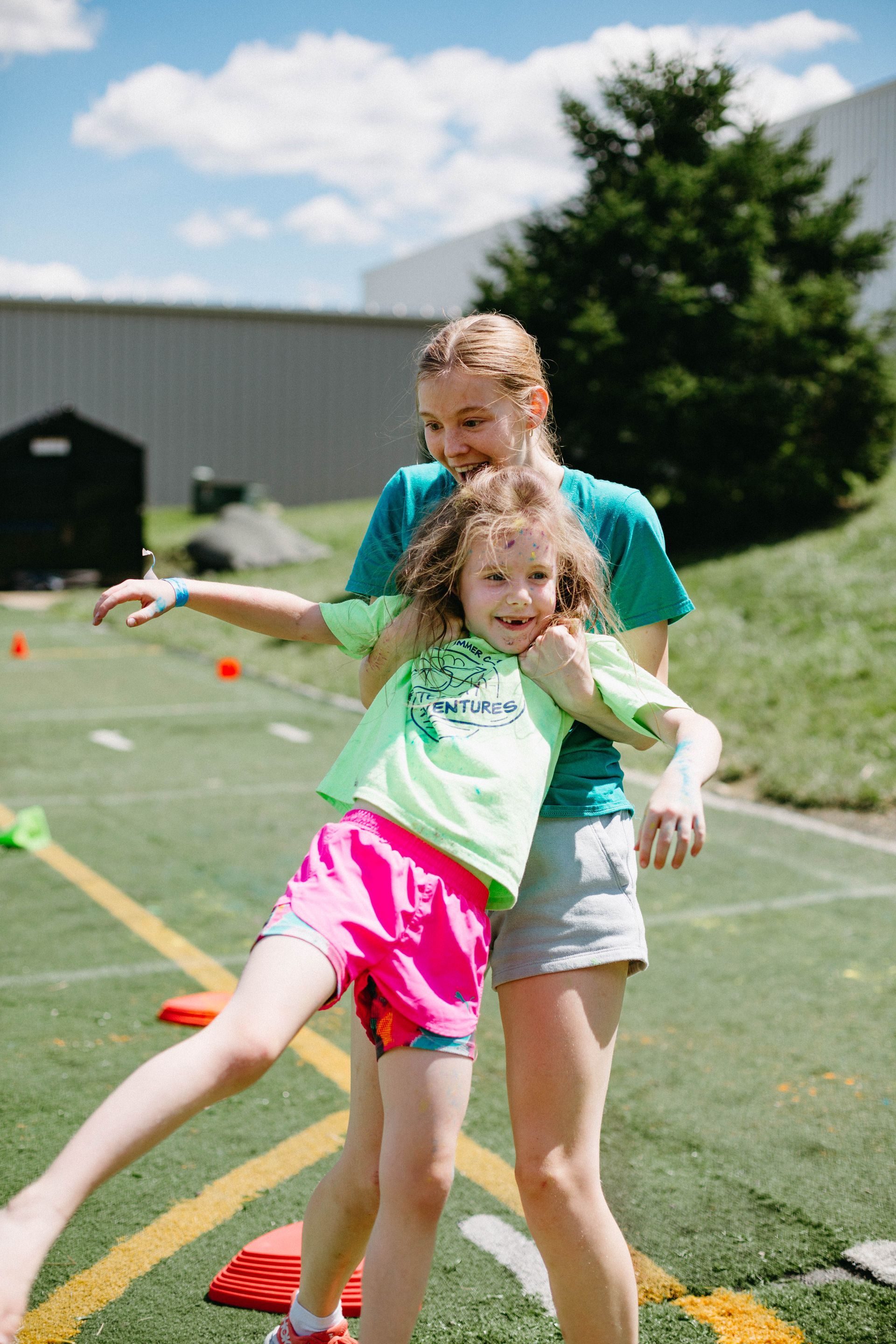 Two children playing outdoors, one balancing another on a sunny grassy field near orange cones.