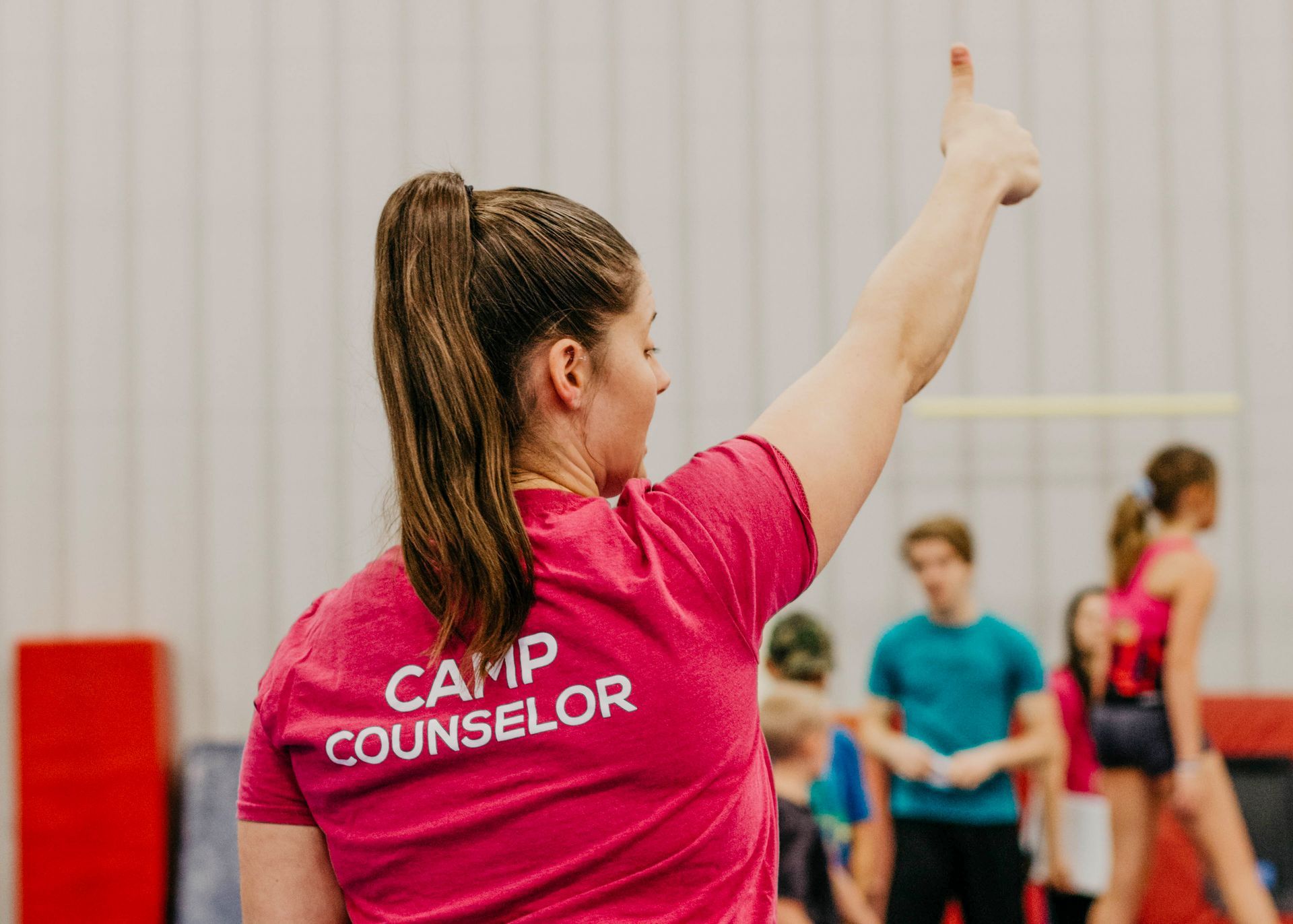 Camp counselor in pink shirt points during an indoor activity with participants in the background