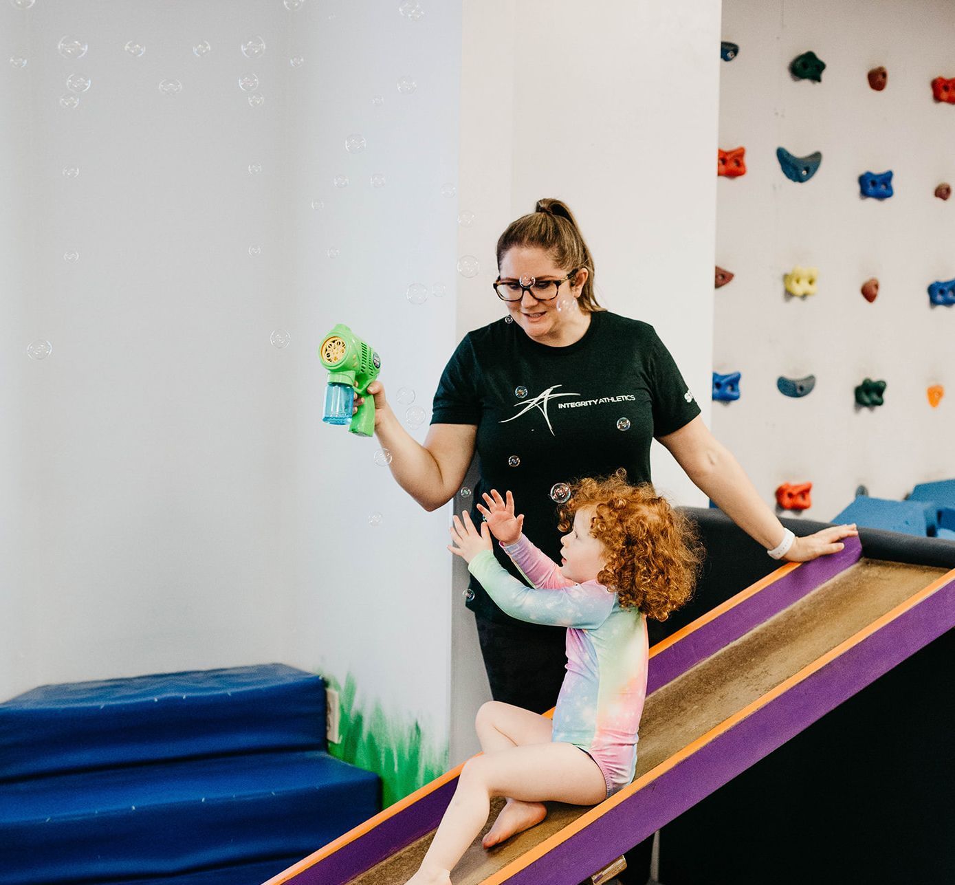 Woman helping child slide indoors while holding a green toy, near a climbing wall