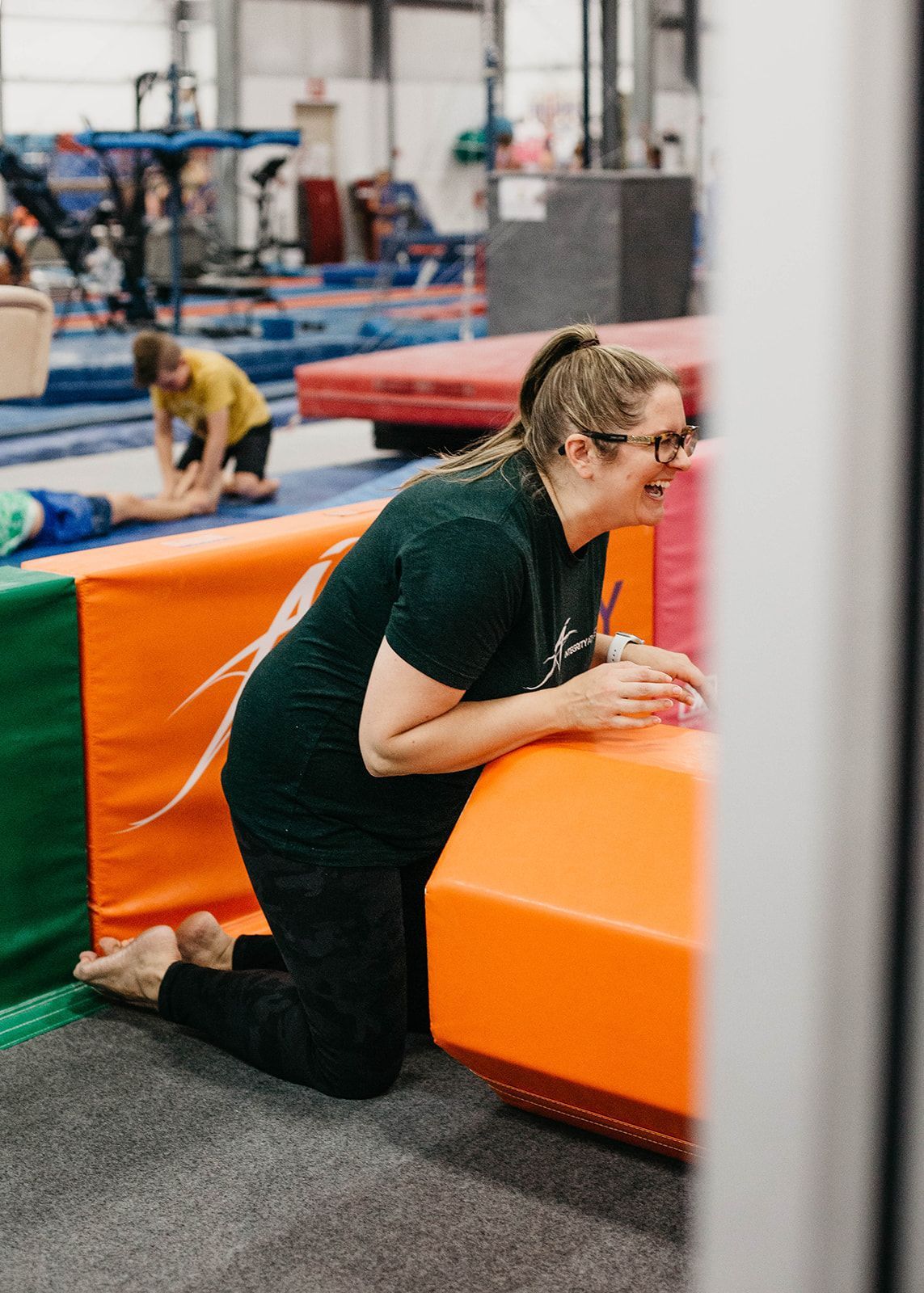 Person kneeling on a gym mat beside an orange foam block in an indoor training area