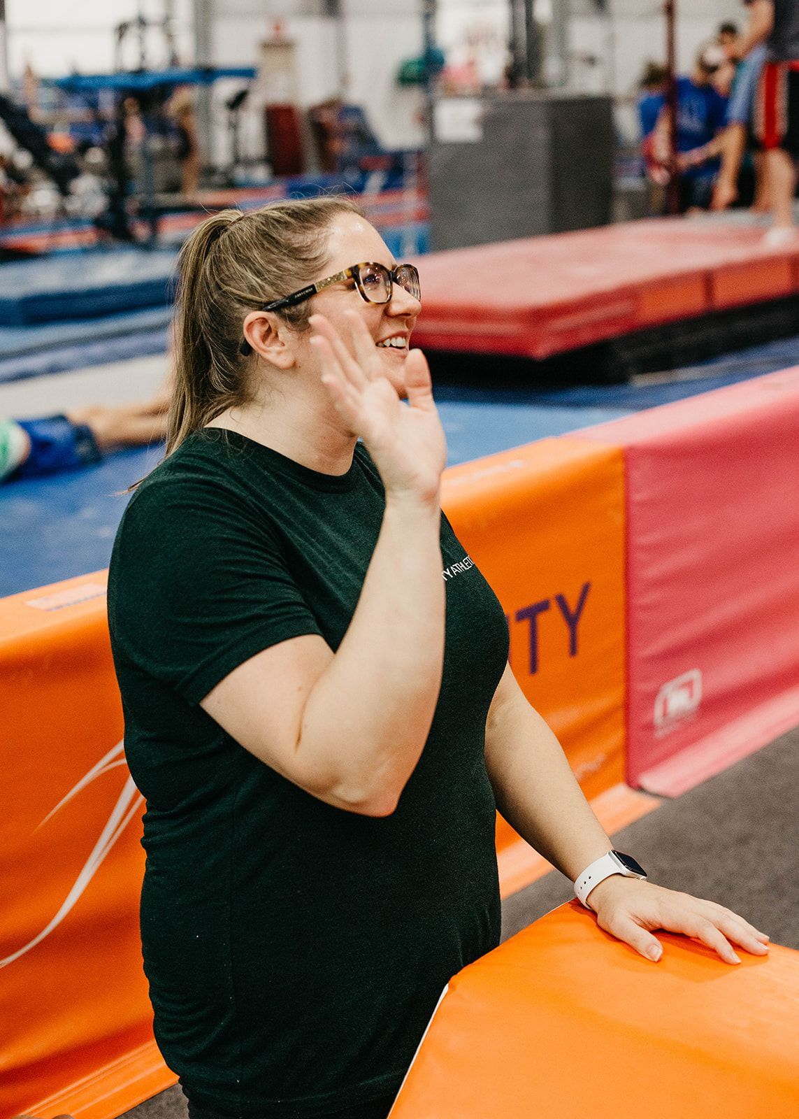 Person in black shirt standing by orange mat at an indoor gymnastics gym, hand raised near face.