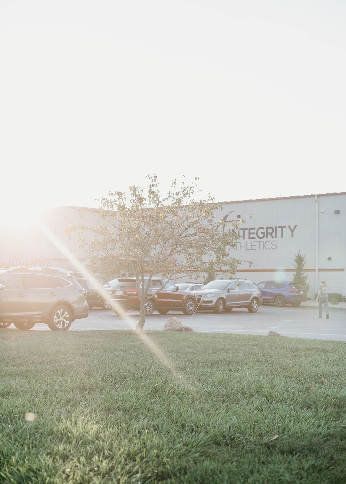 Sunlit parking lot with cars, a tree, and a warehouse labeled “Integrity,” viewed across grass.