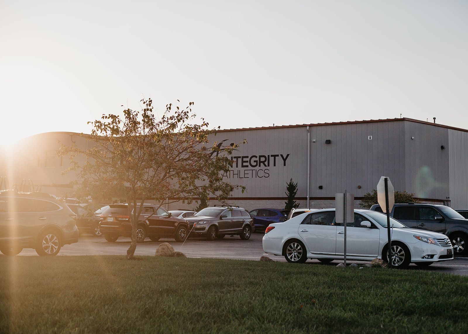 Parking lot outside a gray warehouse at sunset, with cars and a bright sun flare.