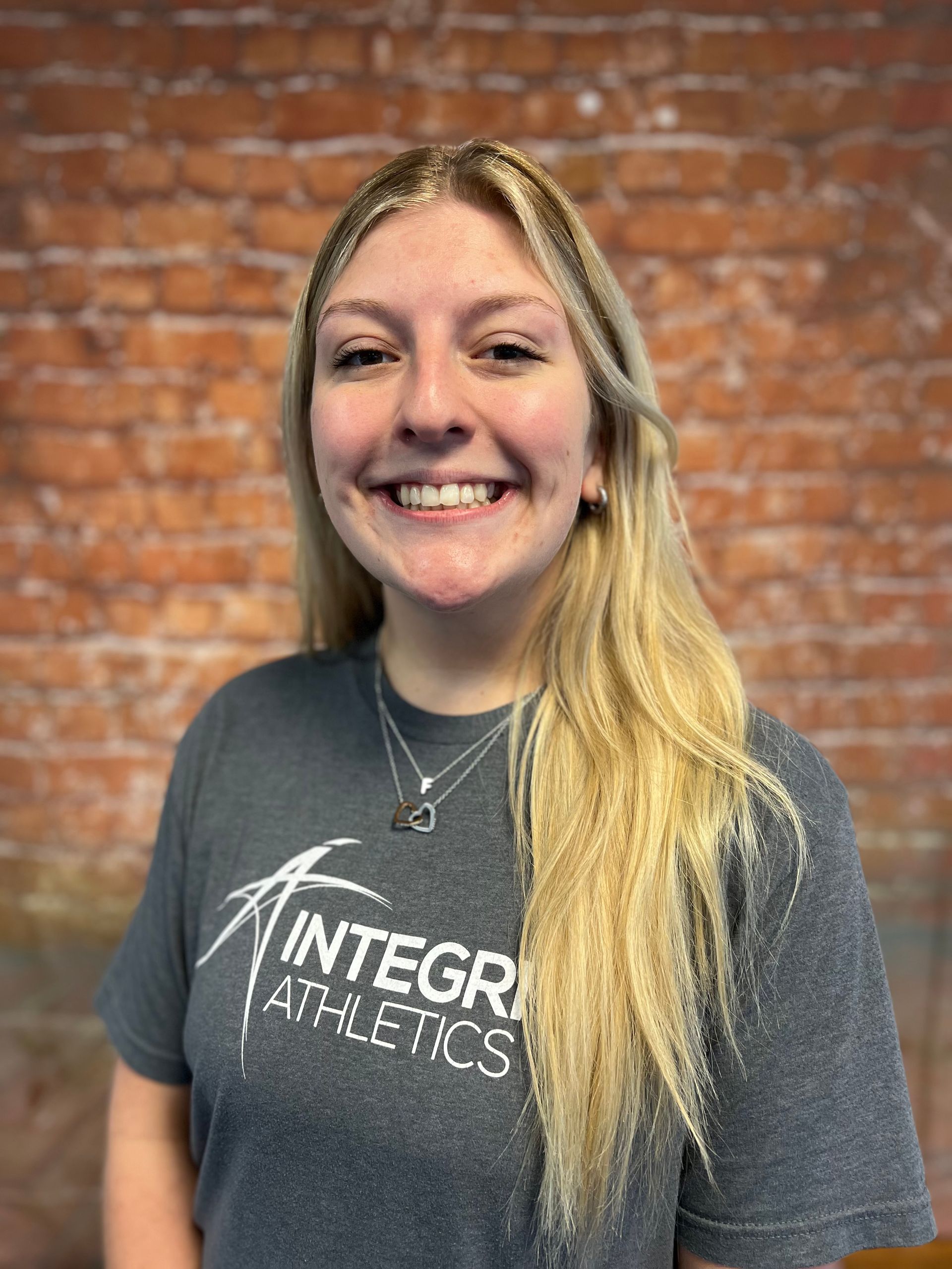 Smiling person in a gray Antigravity Athletics T-shirt standing before a brick wall.