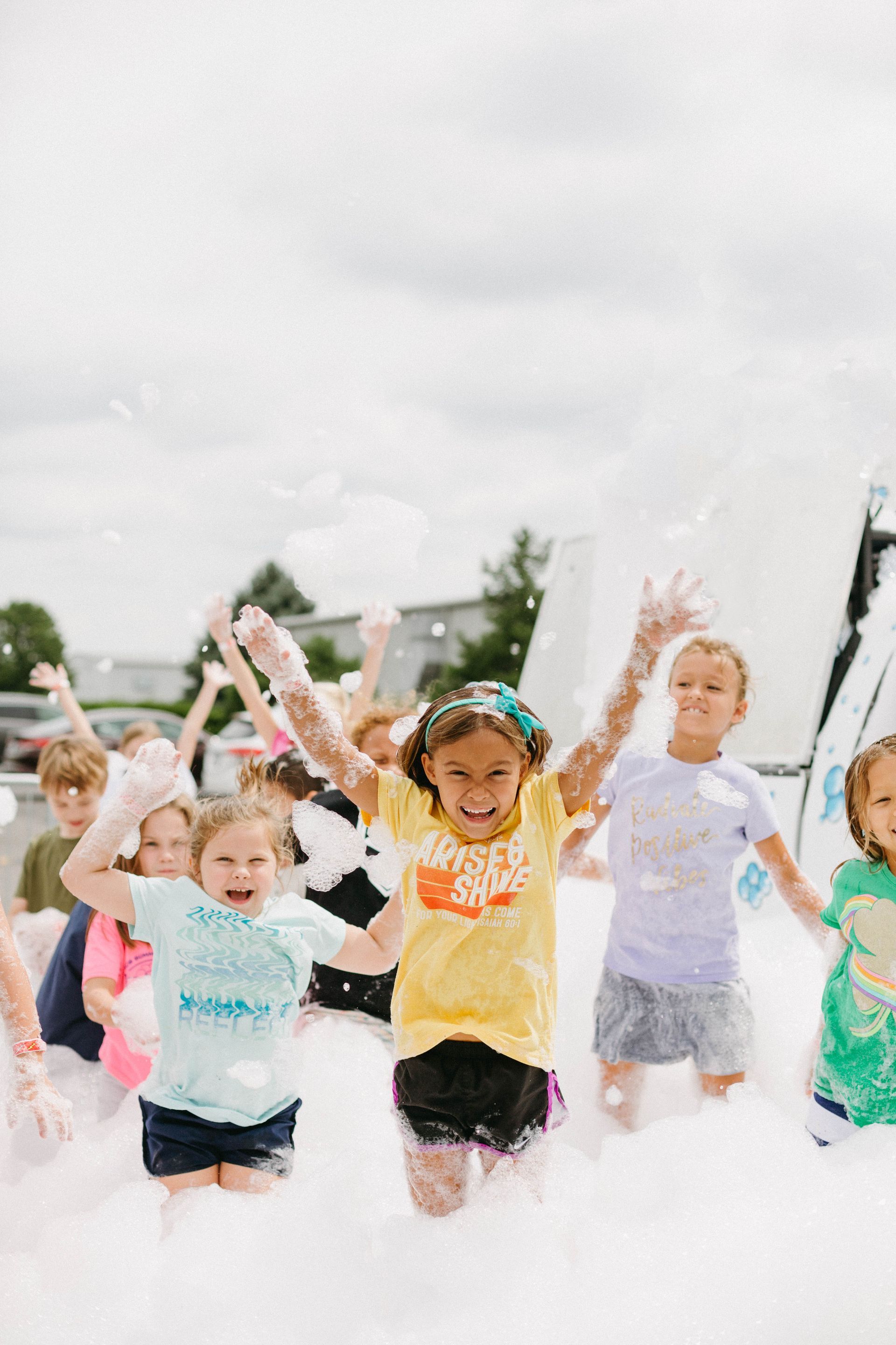 Children running through foam with arms raised at an outdoor event, smiling and cheering