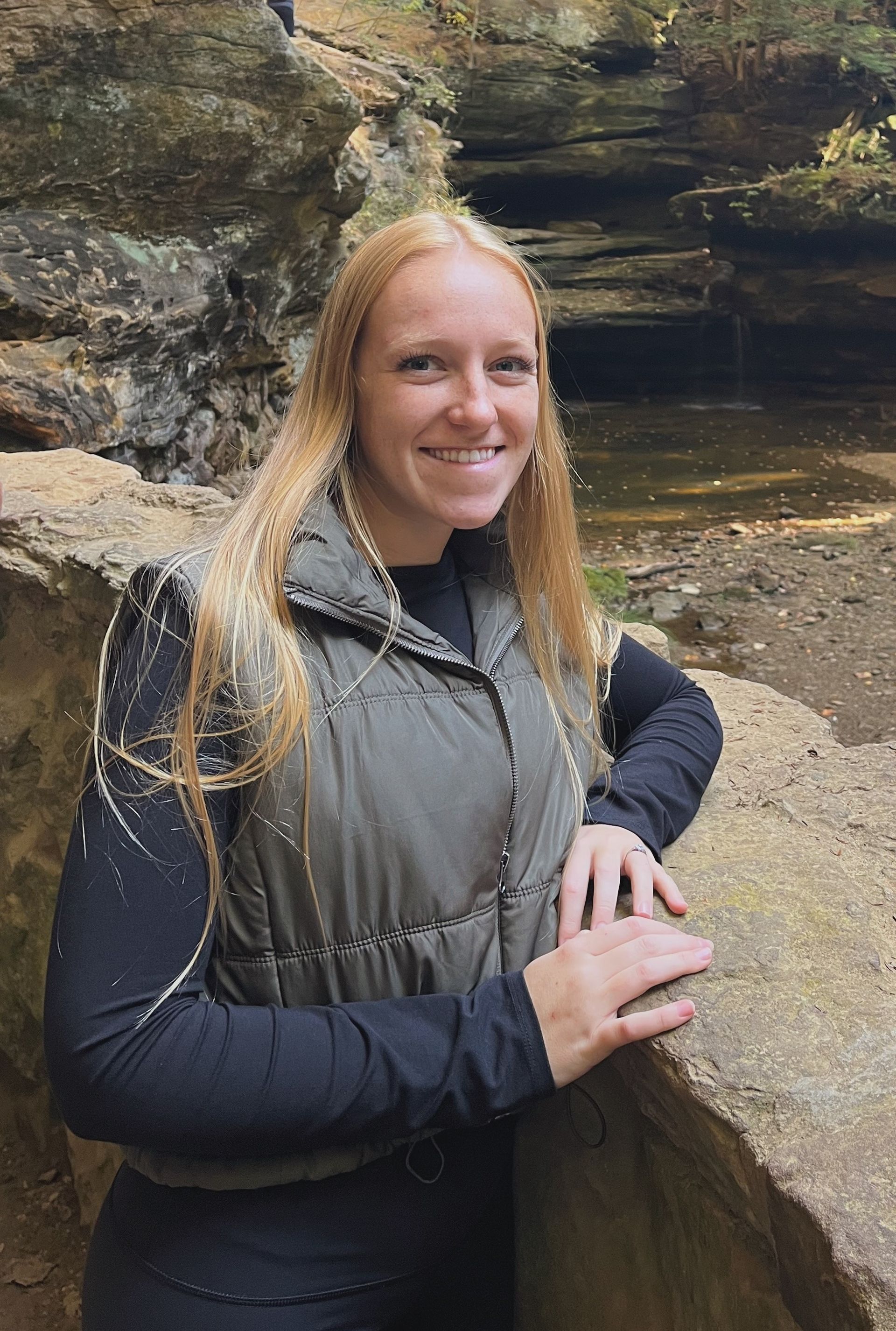 Smiling person in a gray vest stands by a rocky cave wall outdoors