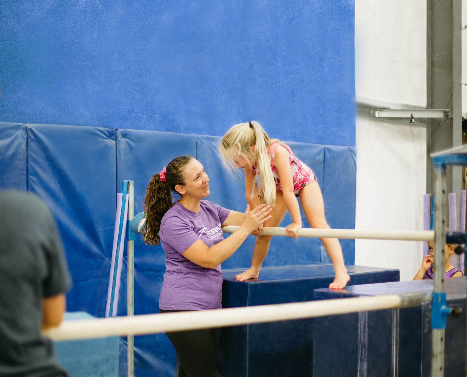 Gymnastics coach helping a child on the uneven bars in a gym with blue mats