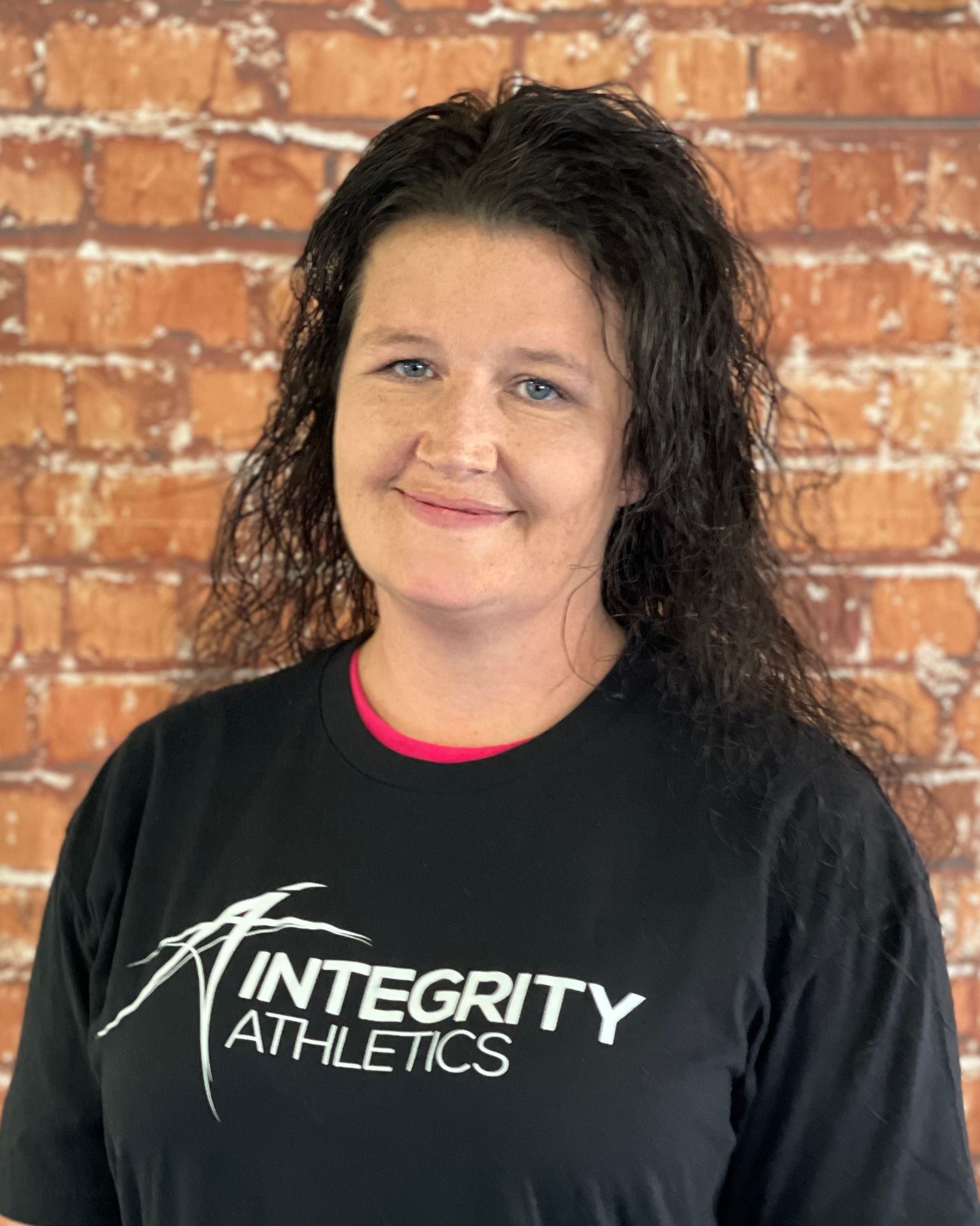Woman in a black Integrity Athletics shirt smiling against a brick wall backdrop