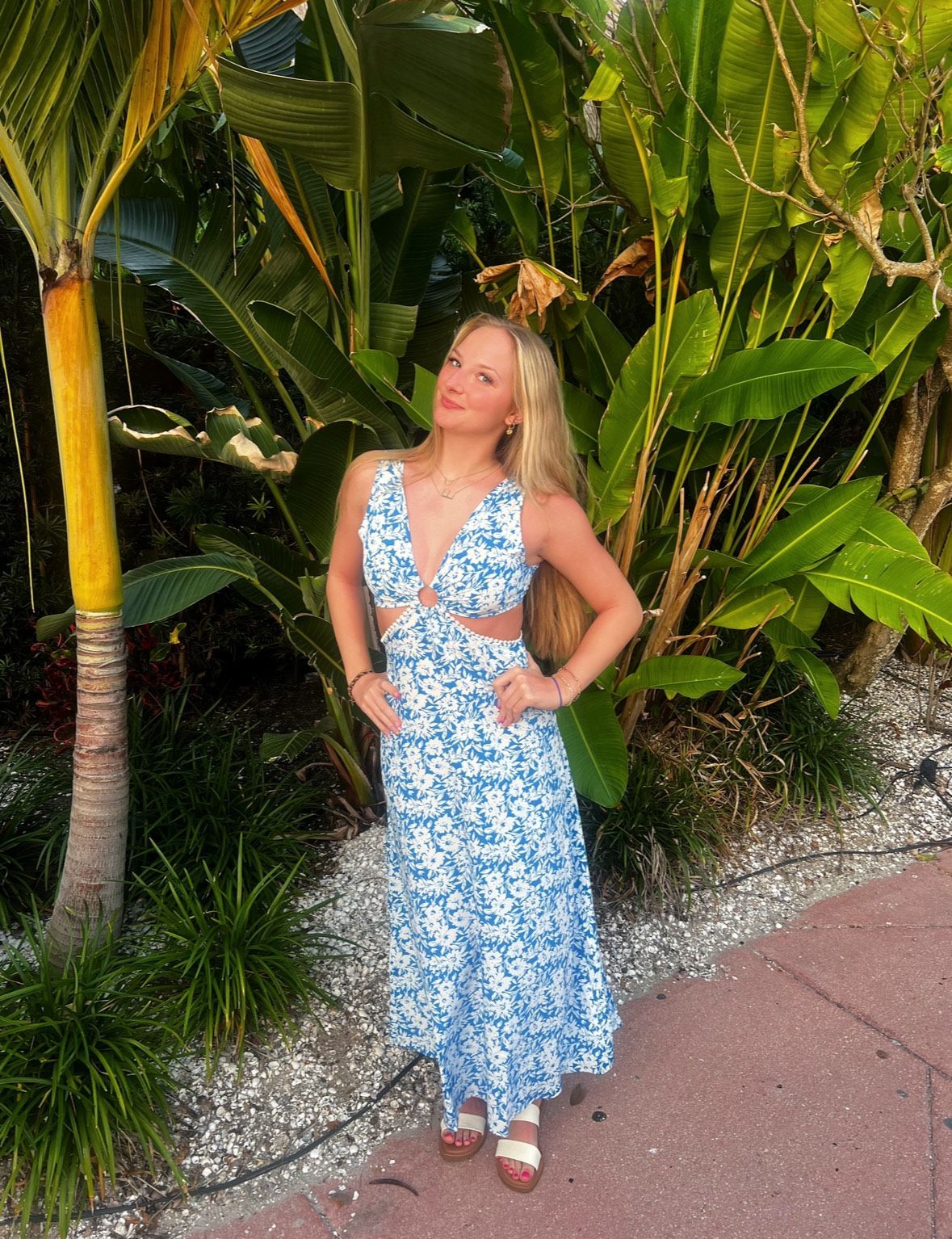 Woman in a blue floral dress standing on a garden path with tropical plants behind her.