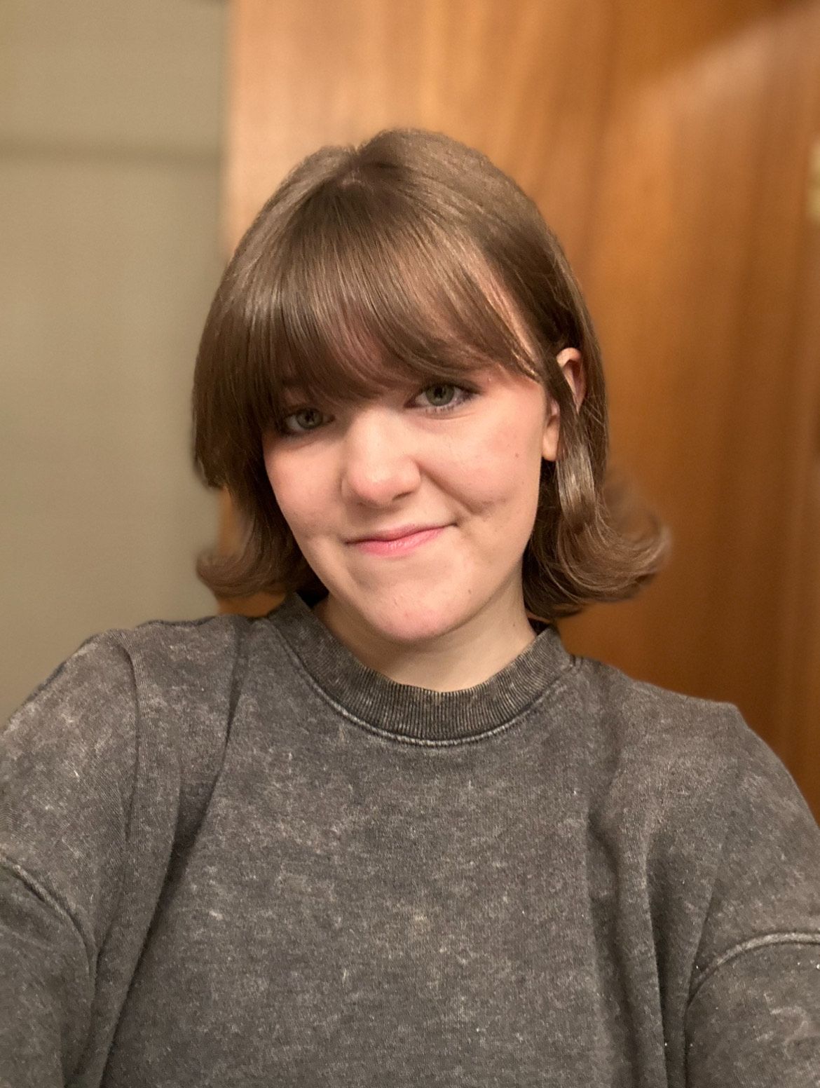 Person in a gray sweater taking a selfie indoors, with short brown hair and a slight smile.