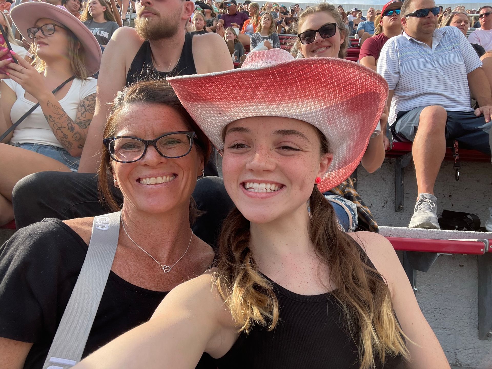 Two smiling people take a selfie at a stadium, one wearing a red cowboy hat.