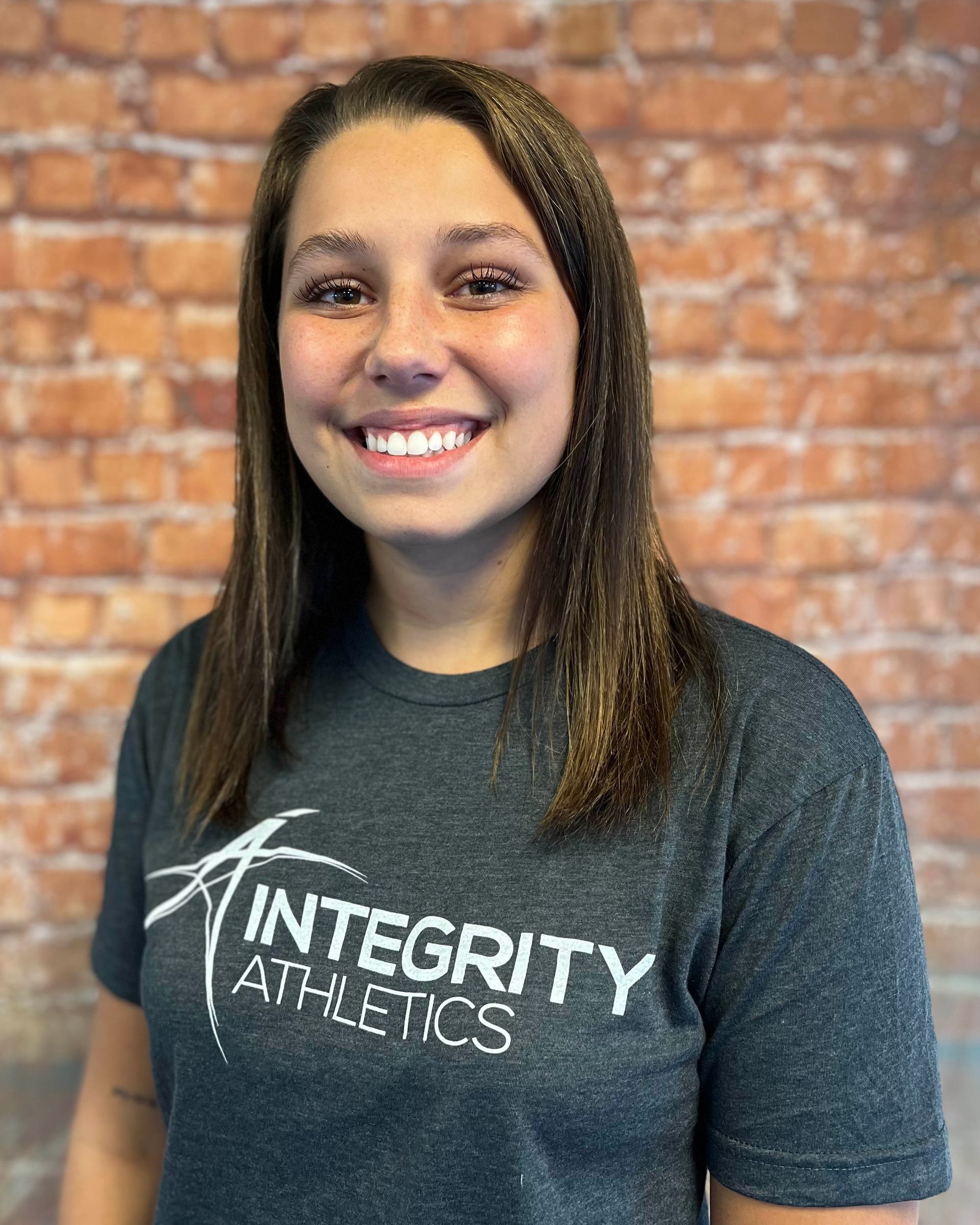 Smiling person in a dark Integrity Athletics T-shirt in front of a brick wall