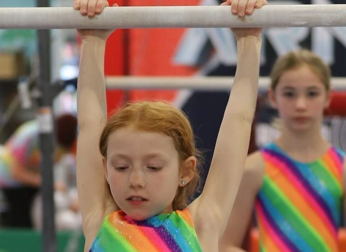 A little girl in a rainbow leotard is sitting on a blue ball.