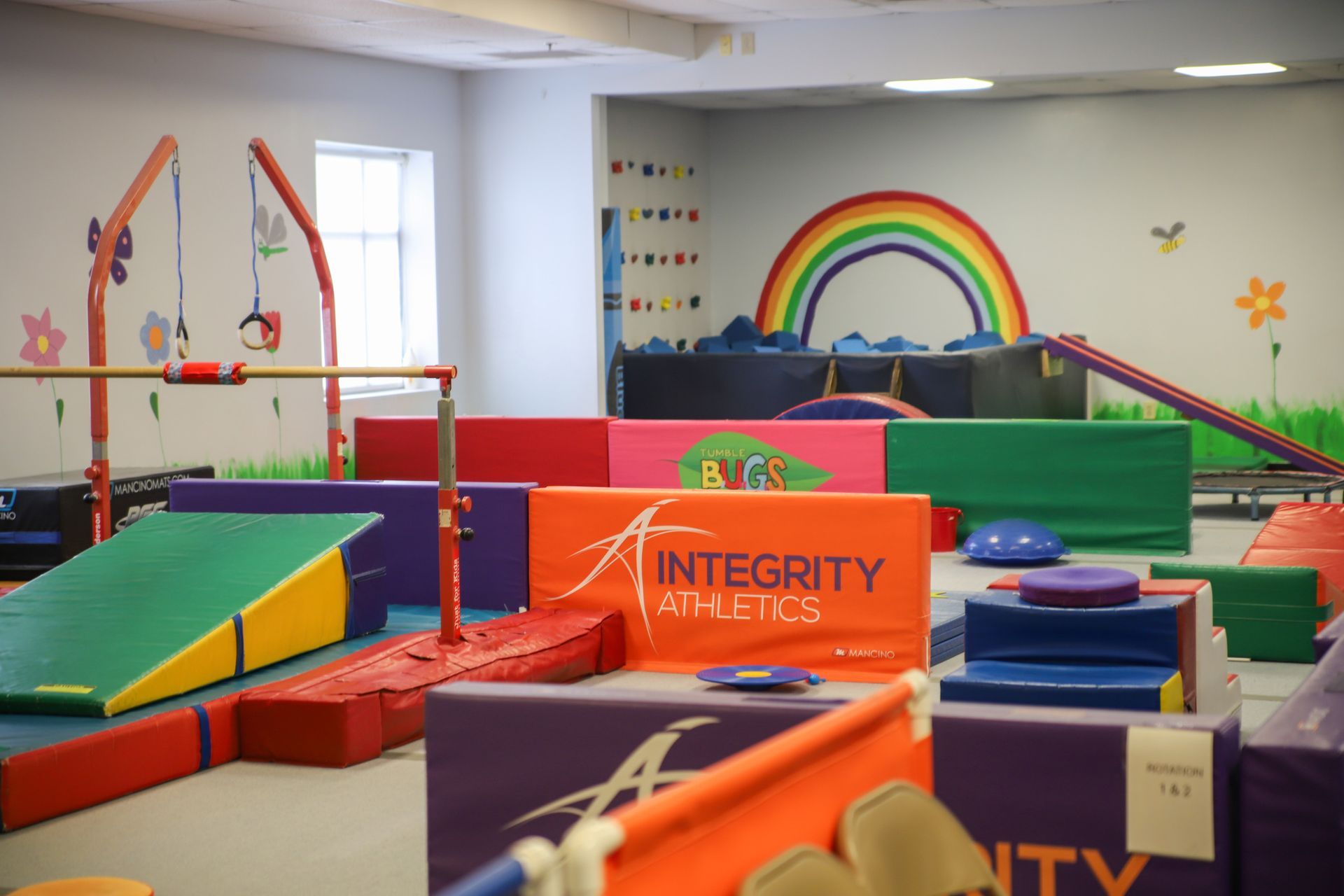 Colorful indoor gymnastics play area with mats, ramps, bars, and a rainbow wall mural