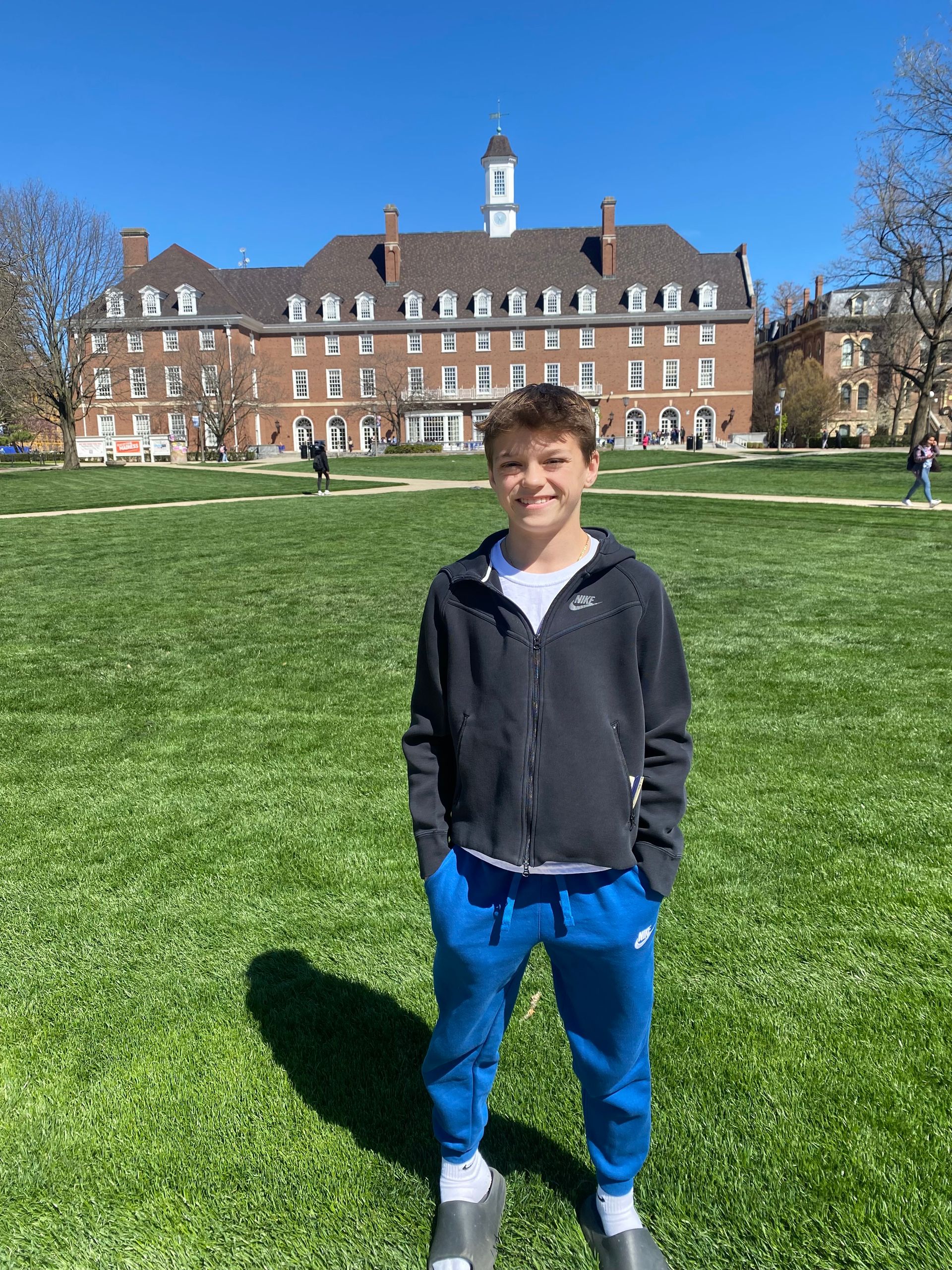 Person standing on a grassy lawn in front of a brick building with a tower under a blue sky