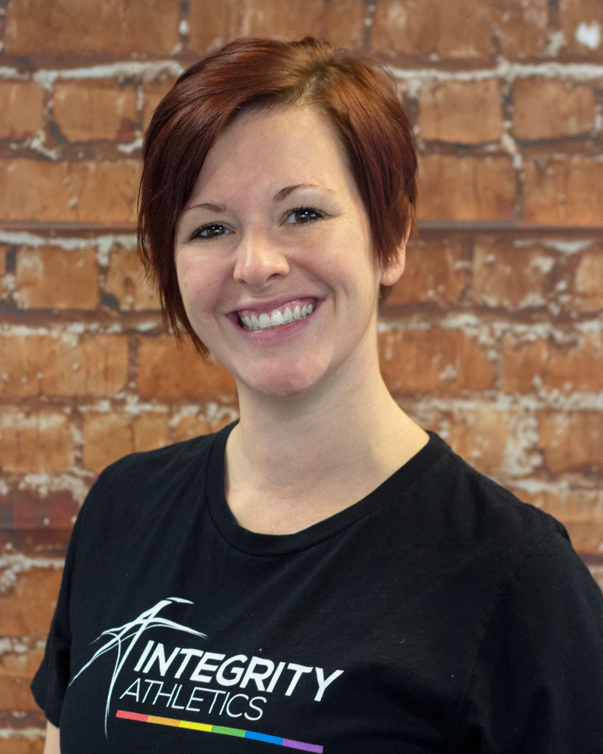Smiling person in a black “Integrity Athletics” shirt standing against a brick wall.