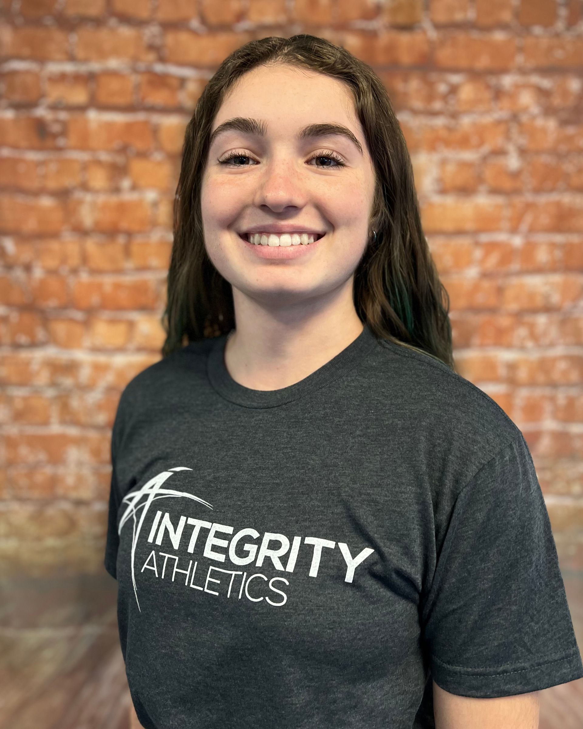 Smiling person in a dark Integrity Athletics T-shirt standing against a brick wall
