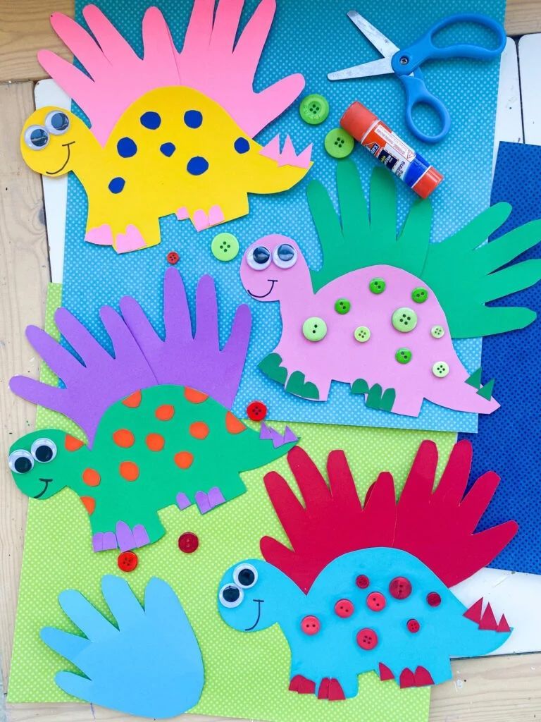 Colorful paper dinosaur craft shapes on a blue background with scissors, glue, and a marker