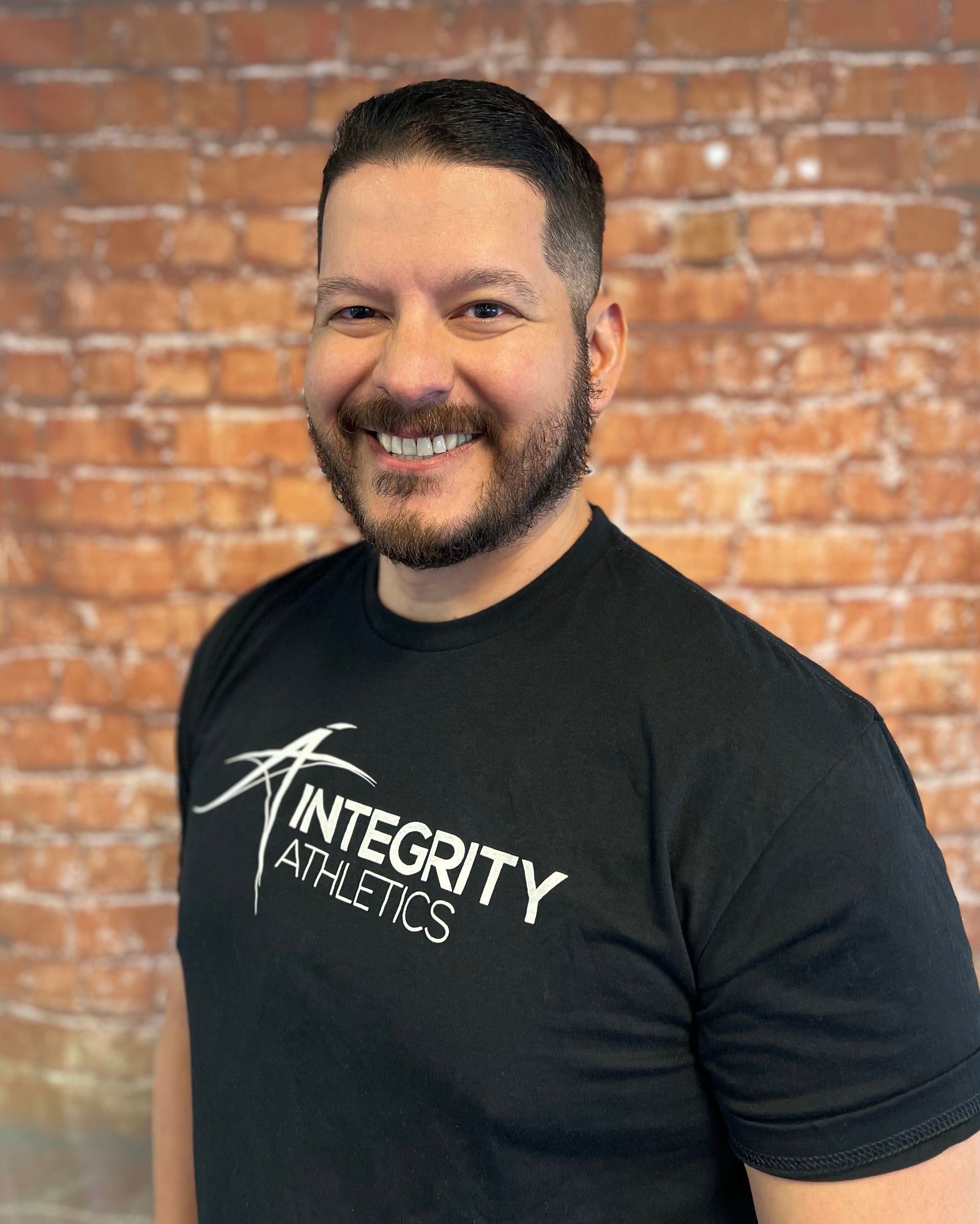 Smiling person in a black “Integrity” T-shirt standing before a brick wall.