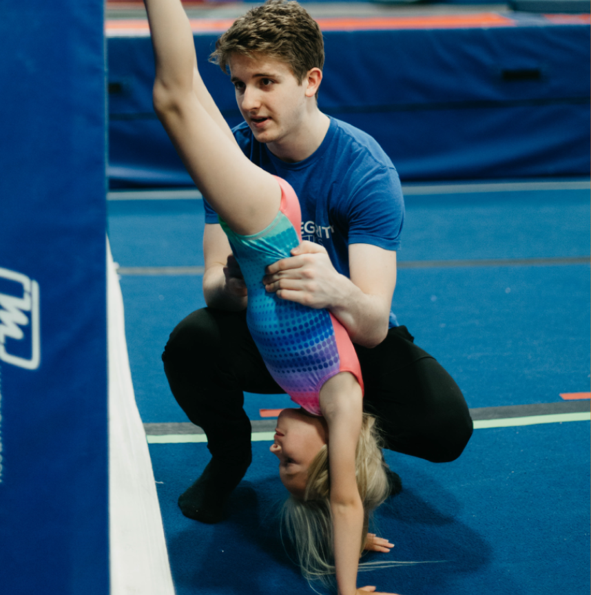 A man is helping a young girl do a handstand on a blue mat