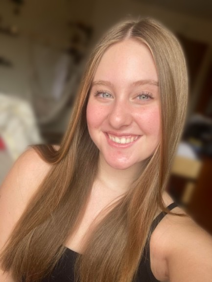 Smiling woman with long blonde hair taking a selfie indoors, wearing a black top.