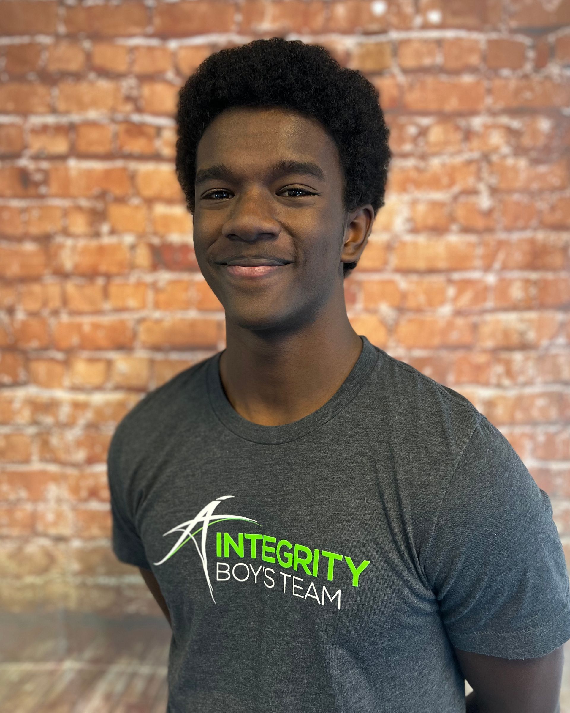 Smiling person in a gray Integrity Boys Team T-shirt against a brick wall backdrop