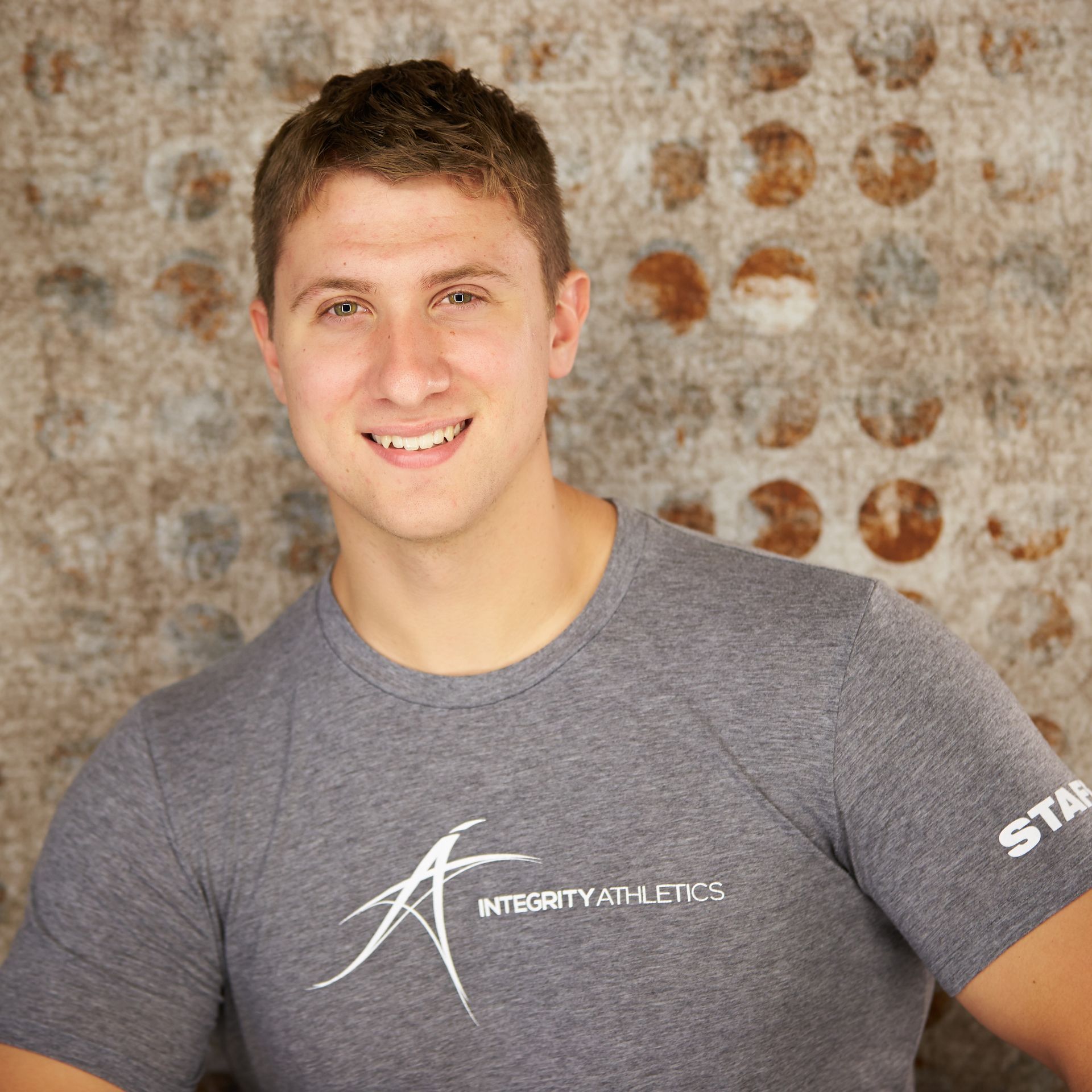 Smiling person in a gray T-shirt standing against a textured wall with rust-colored circles