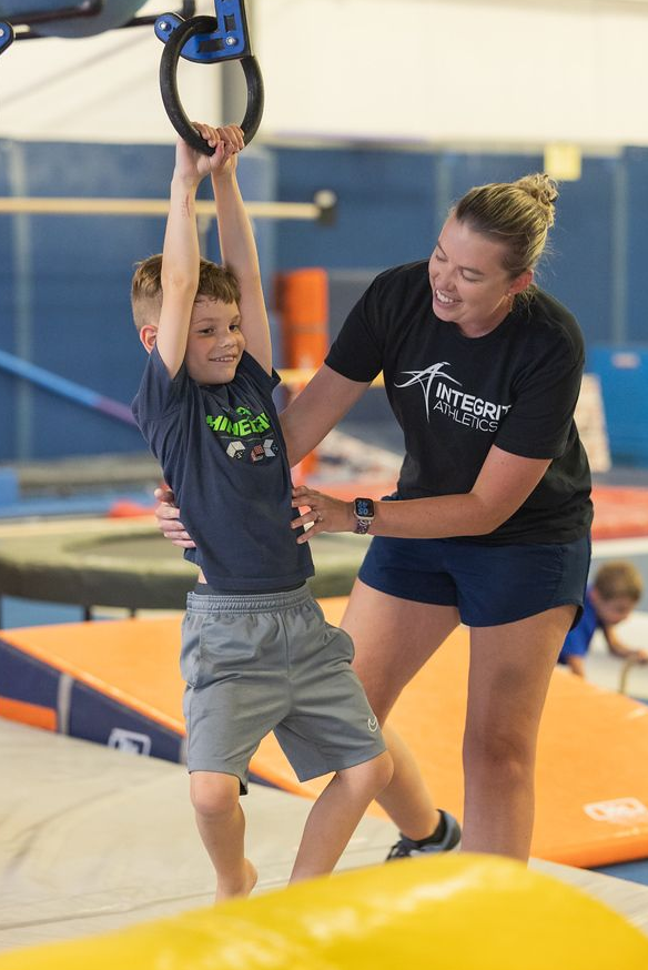 Child hanging from gym rings while a trainer assists on a padded obstacle course