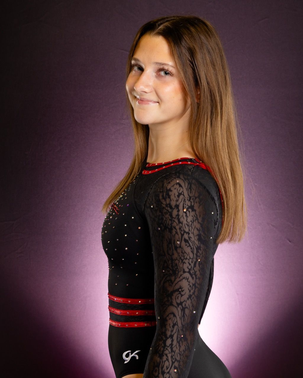 Woman in a black lace dress with red trim, posing against a purple backdrop, smiling over her shoulder