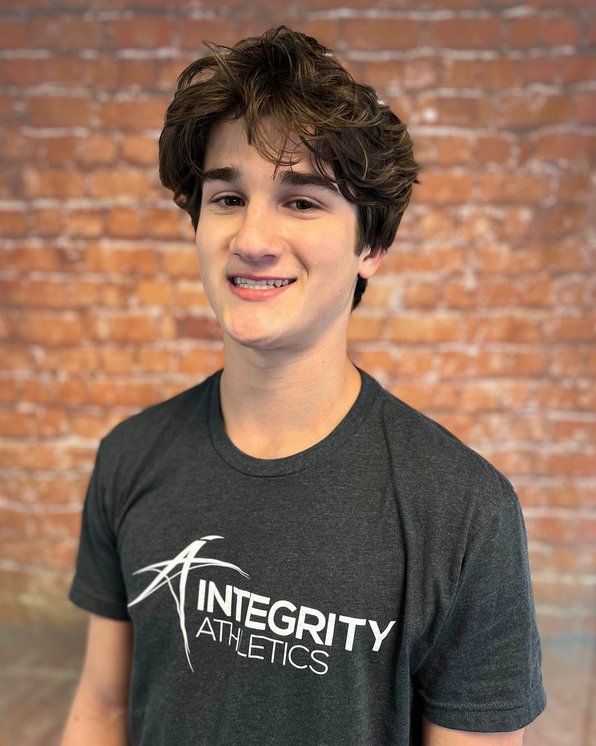 Young person smiling in a dark Integrity Athletics T-shirt against a brick wall background